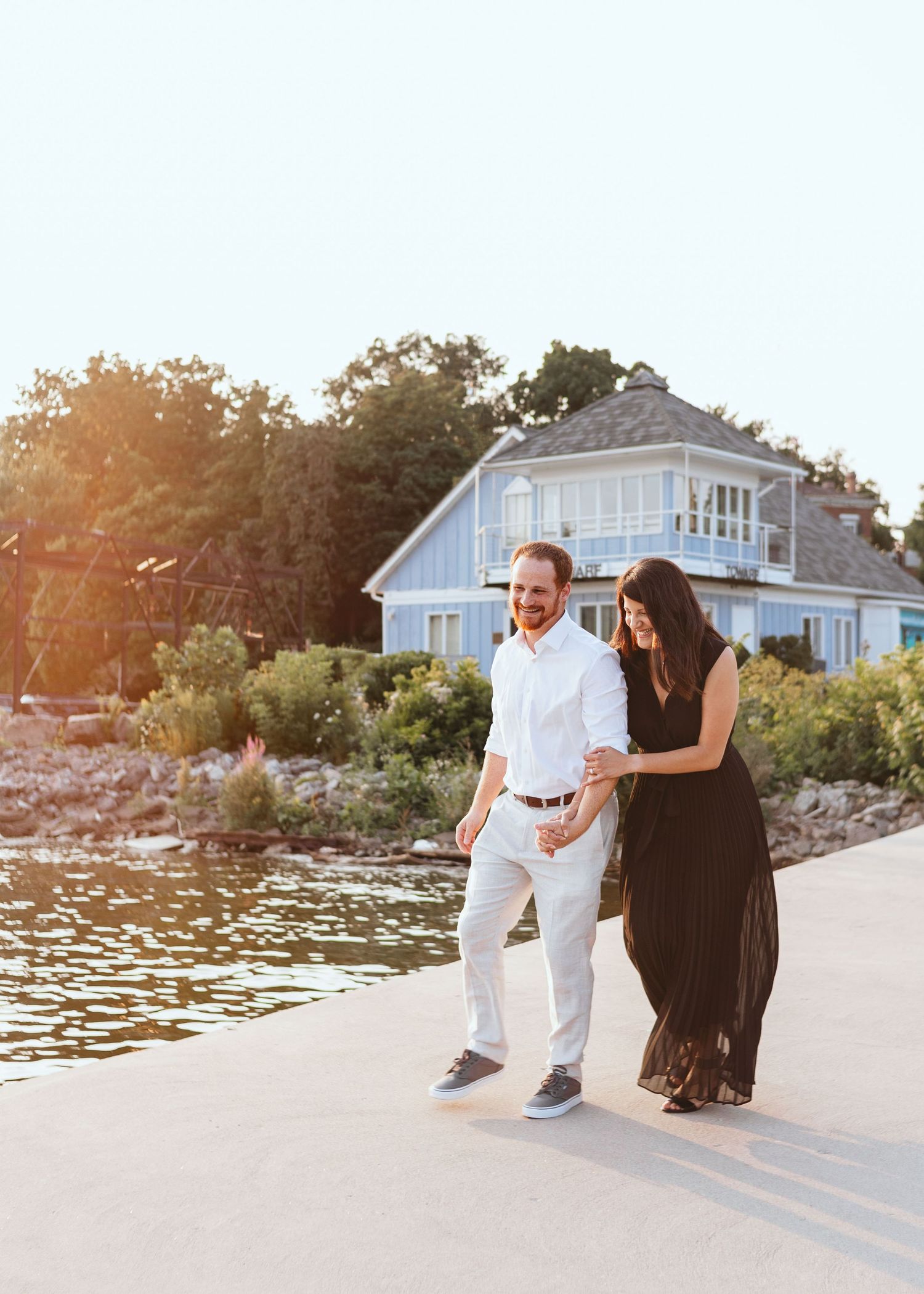A couple walks along a waterfront path at sunset near a coastal home.