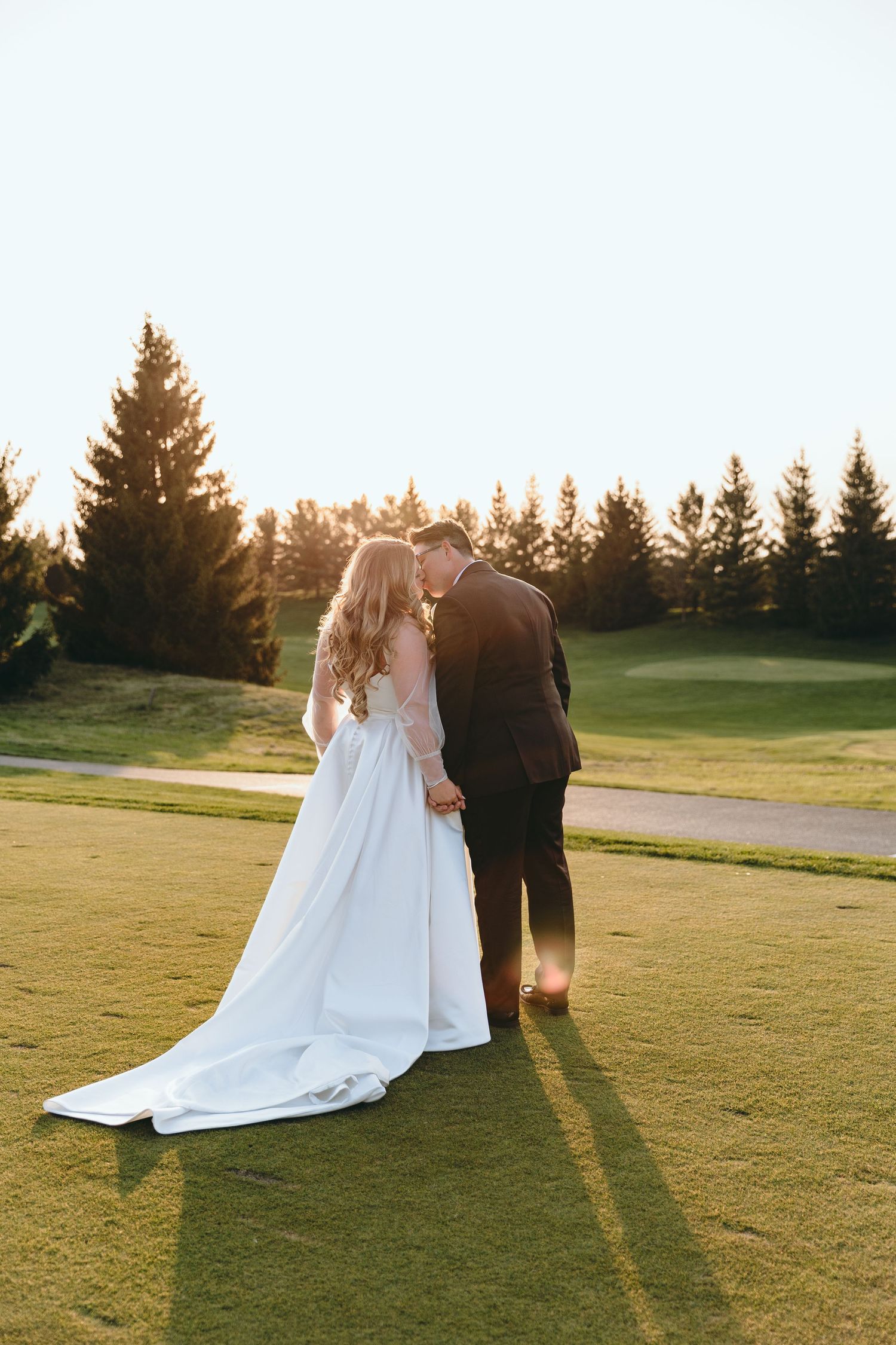 Newlyweds walk hand in hand on a golf course at sunset with pine trees in the background.