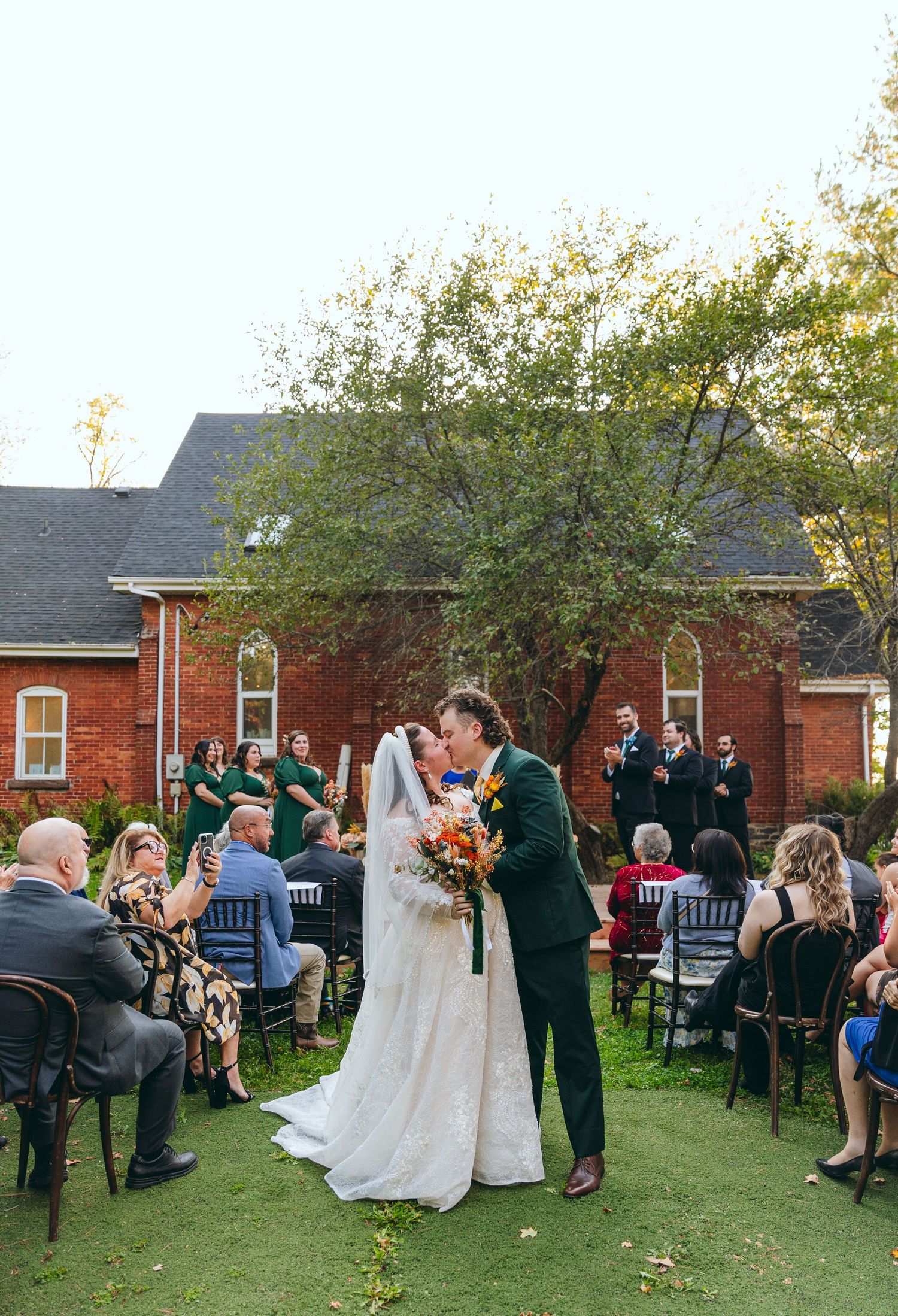 Outdoor wedding ceremony takes place on green lawn with red brick building in background.