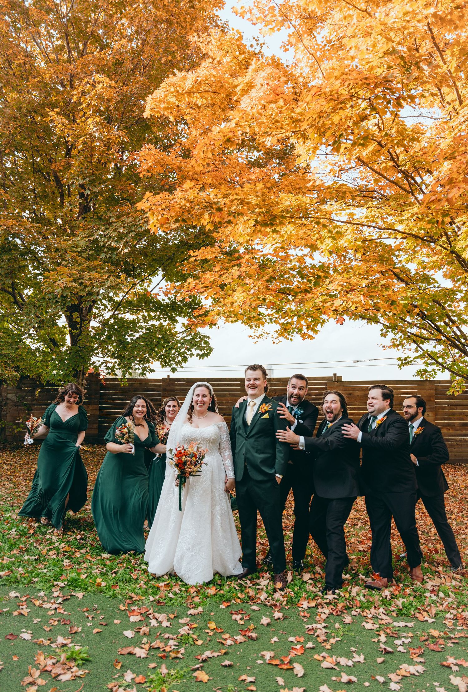 Wedding party in emerald green and black attire poses under vibrant fall foliage.