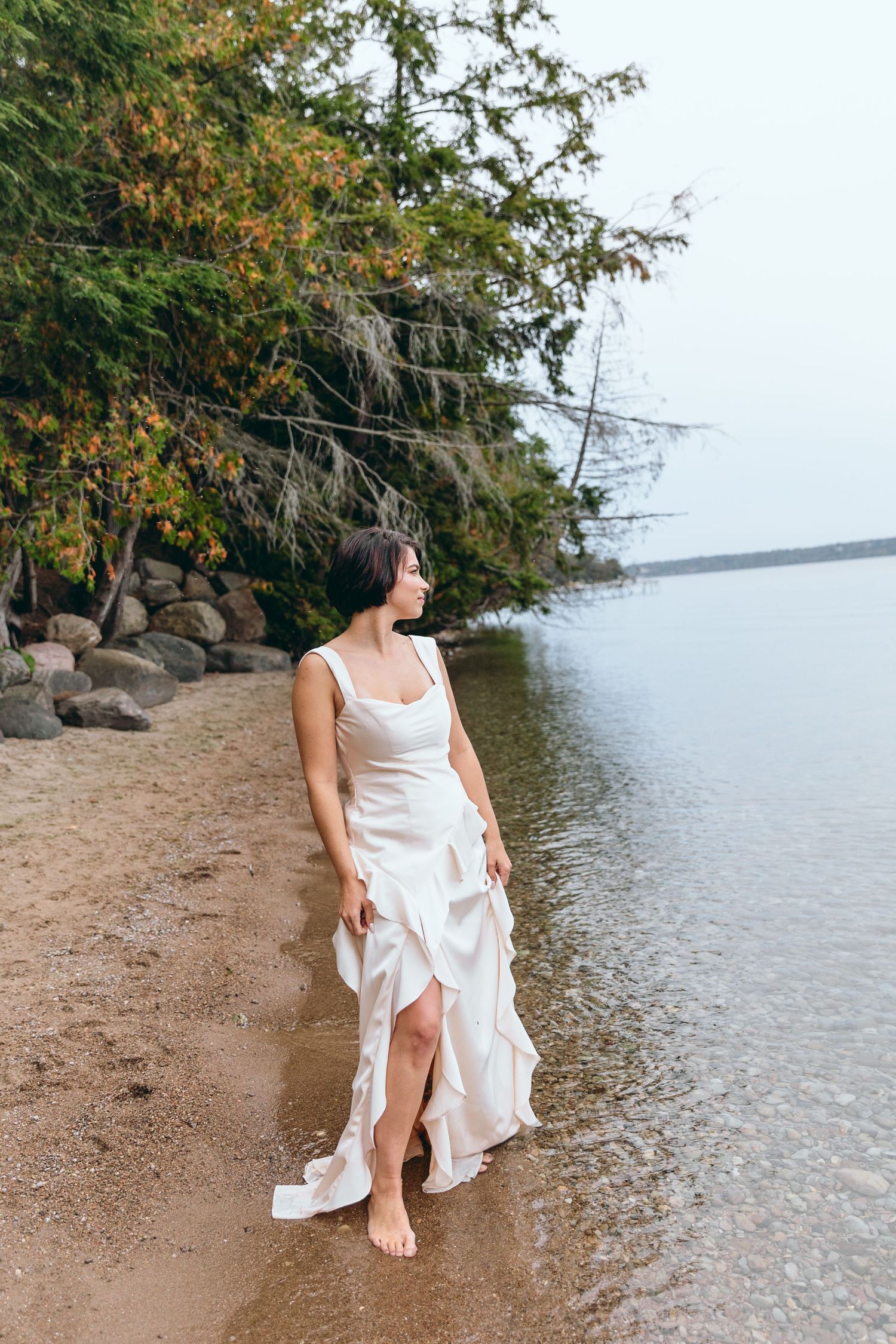 Woman in flowing white dress standing at water's edge on beach.