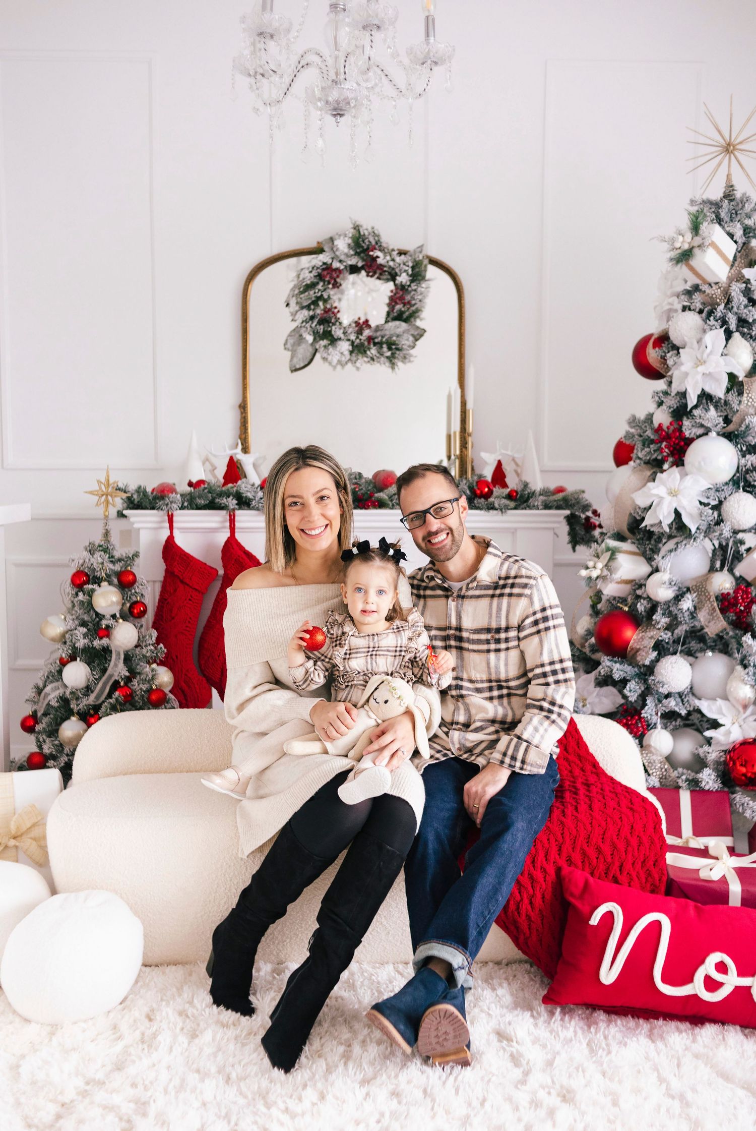 Family of three posing for Christmas photos with decorated trees and stockings in a festive white studio setting.