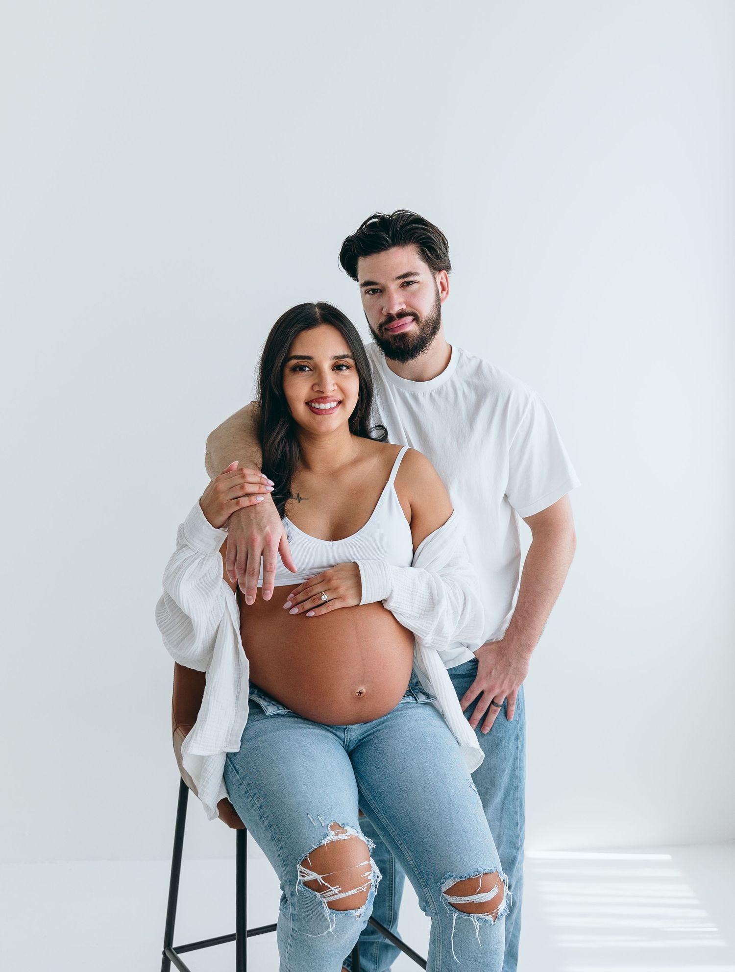 Couple's maternity photoshoot in casual outfits against white backdrop.