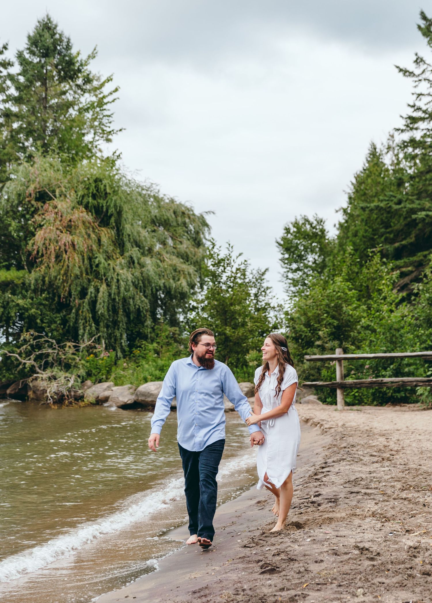 A couple walks playfully along a beach shoreline surrounded by evergreen trees.
