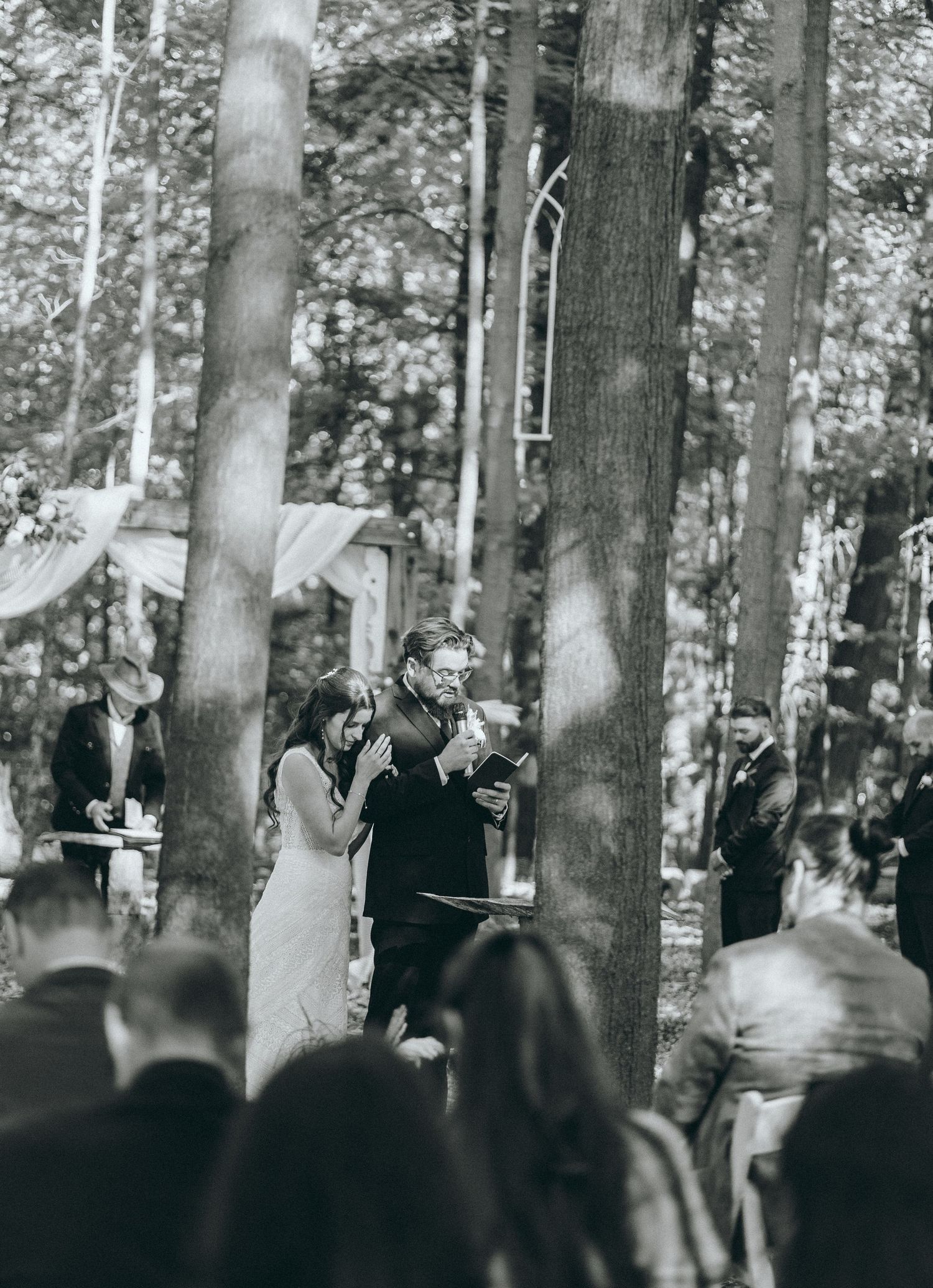 A black and white capture of a woodland wedding ceremony with guests watching from wooden chairs.