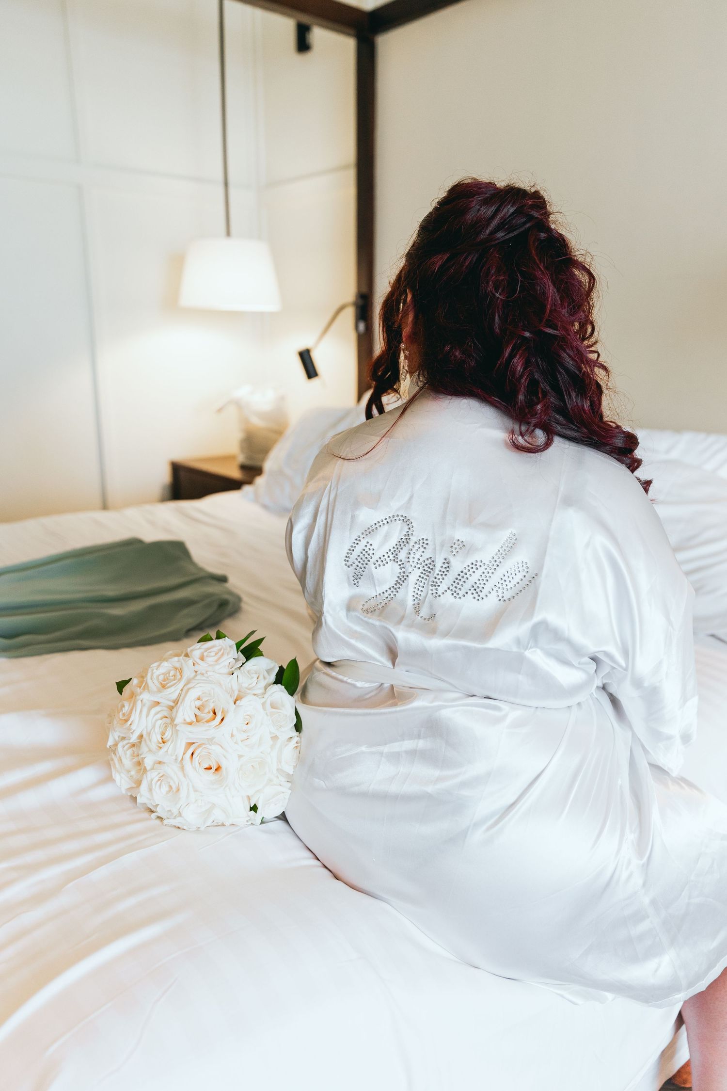 White bridal robe with text lies on bed next to delicate white roses in wedding preparation setting.