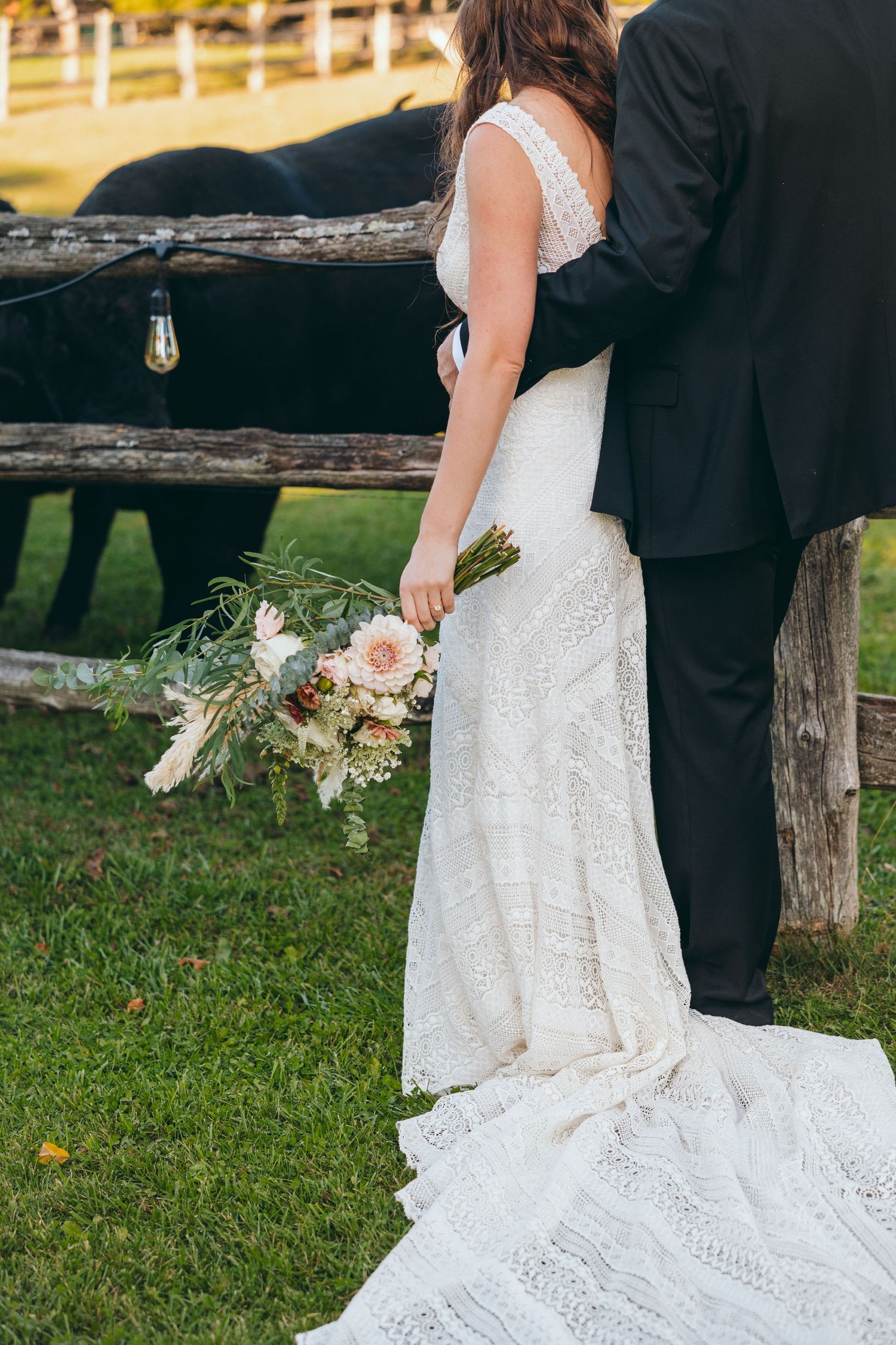 Wedding couple poses near rustic wooden fence with flowing white dress and bouquet.