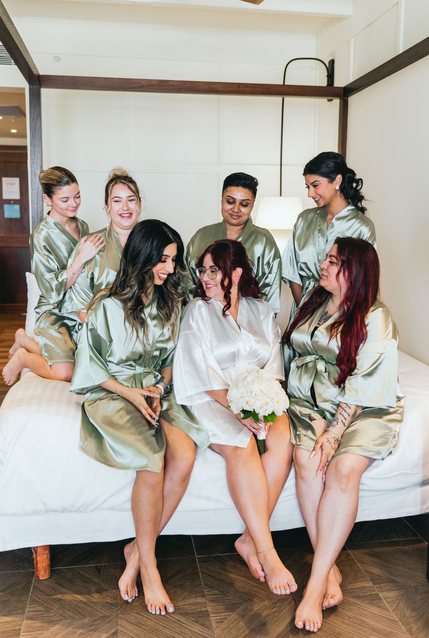 Bridesmaids in sage green silk robes sit together on a bed sharing laughter during wedding preparations.