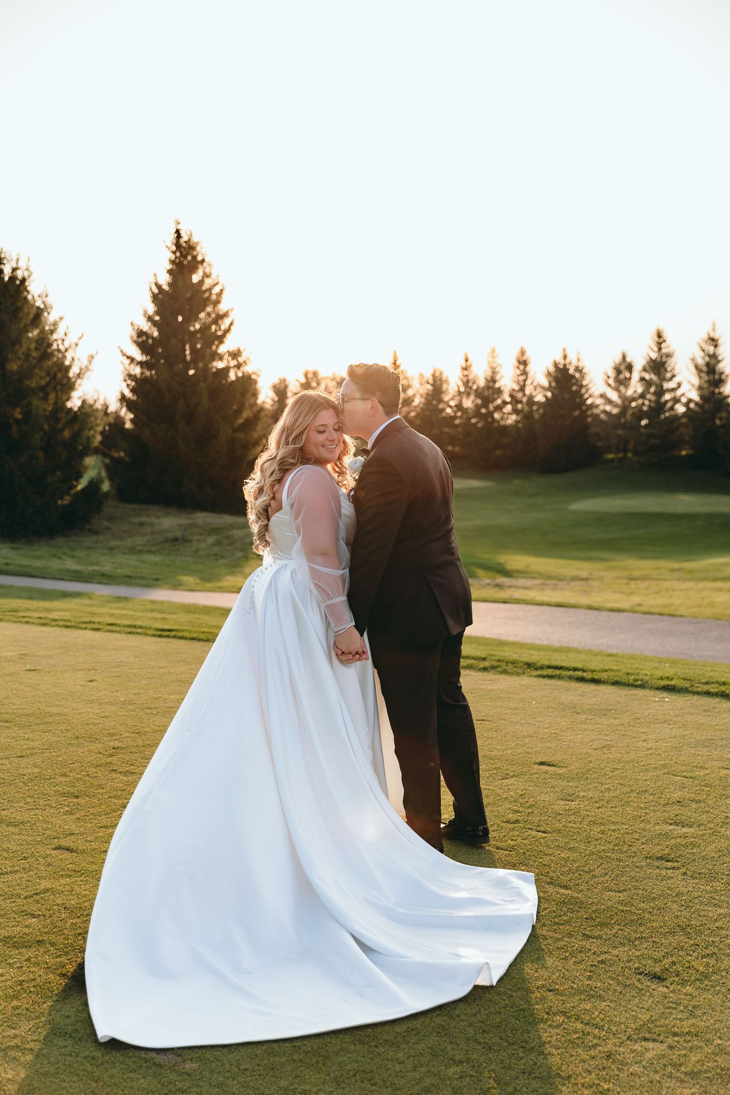 Golden sunset light illuminates a couple in wedding attire on a manicured lawn.