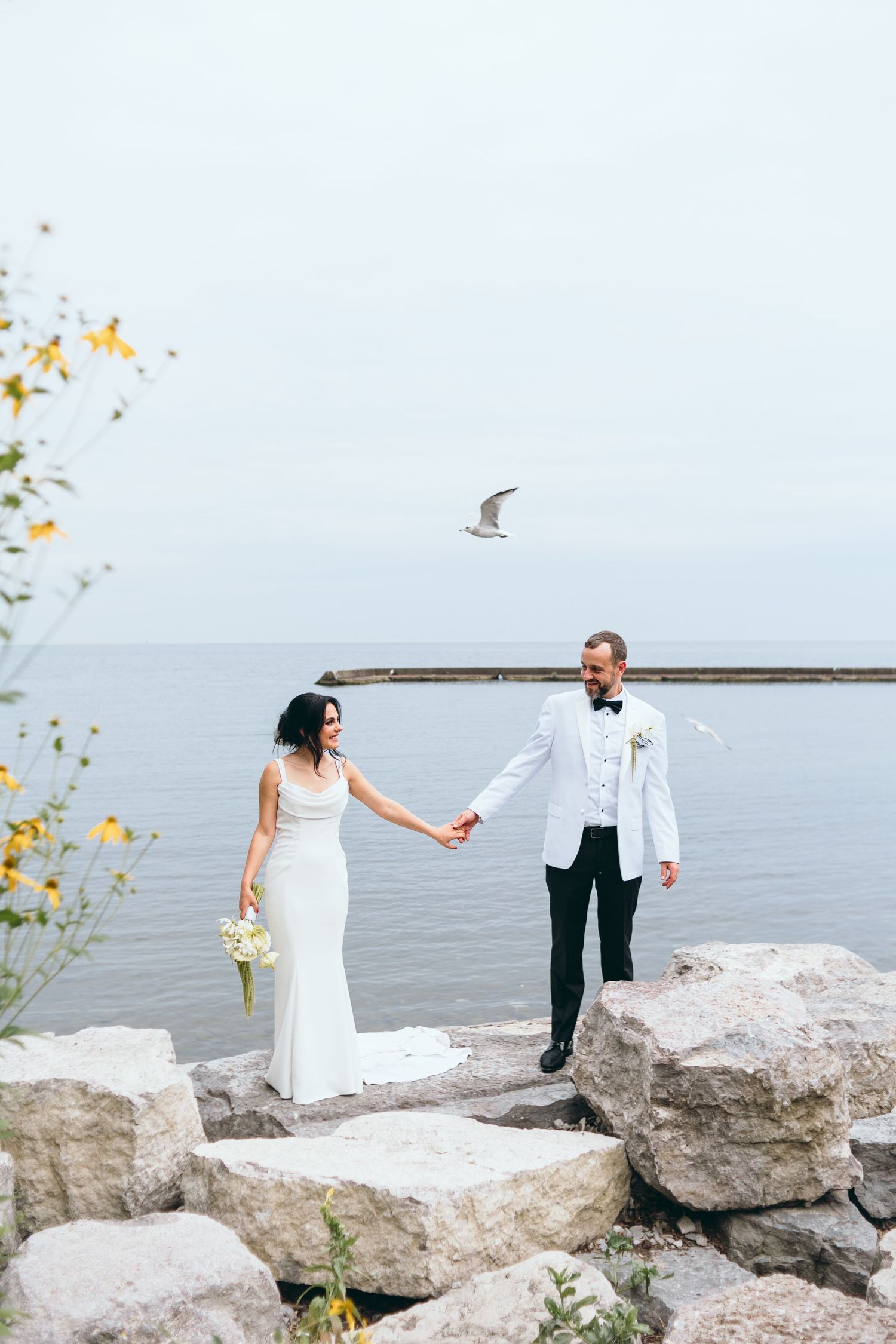 Newlyweds pose romantically on rocky shoreline with ocean and seagulls in background.