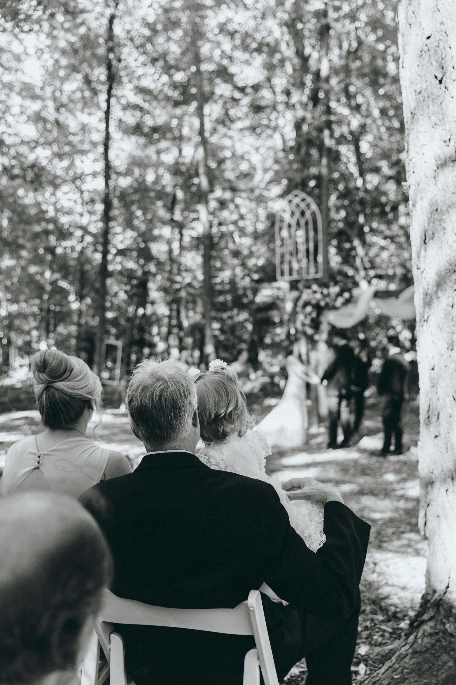 Black and white image of wedding guests watching ceremony in wooded outdoor venue.