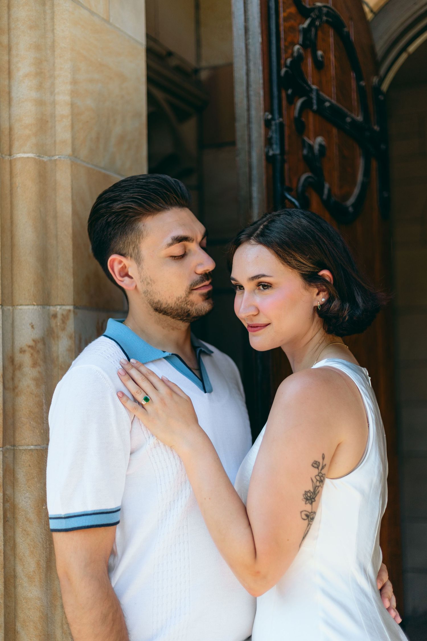 A romantic couple embraces against a stone wall with ornate architectural details in soft lighting.