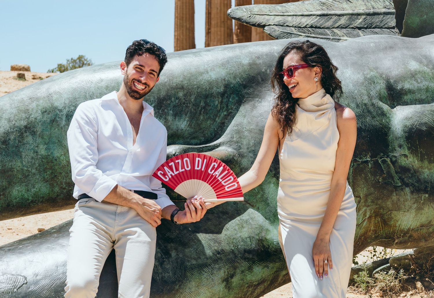 Playful couple poses near large fallen statue at ancient Greek archaeological site.