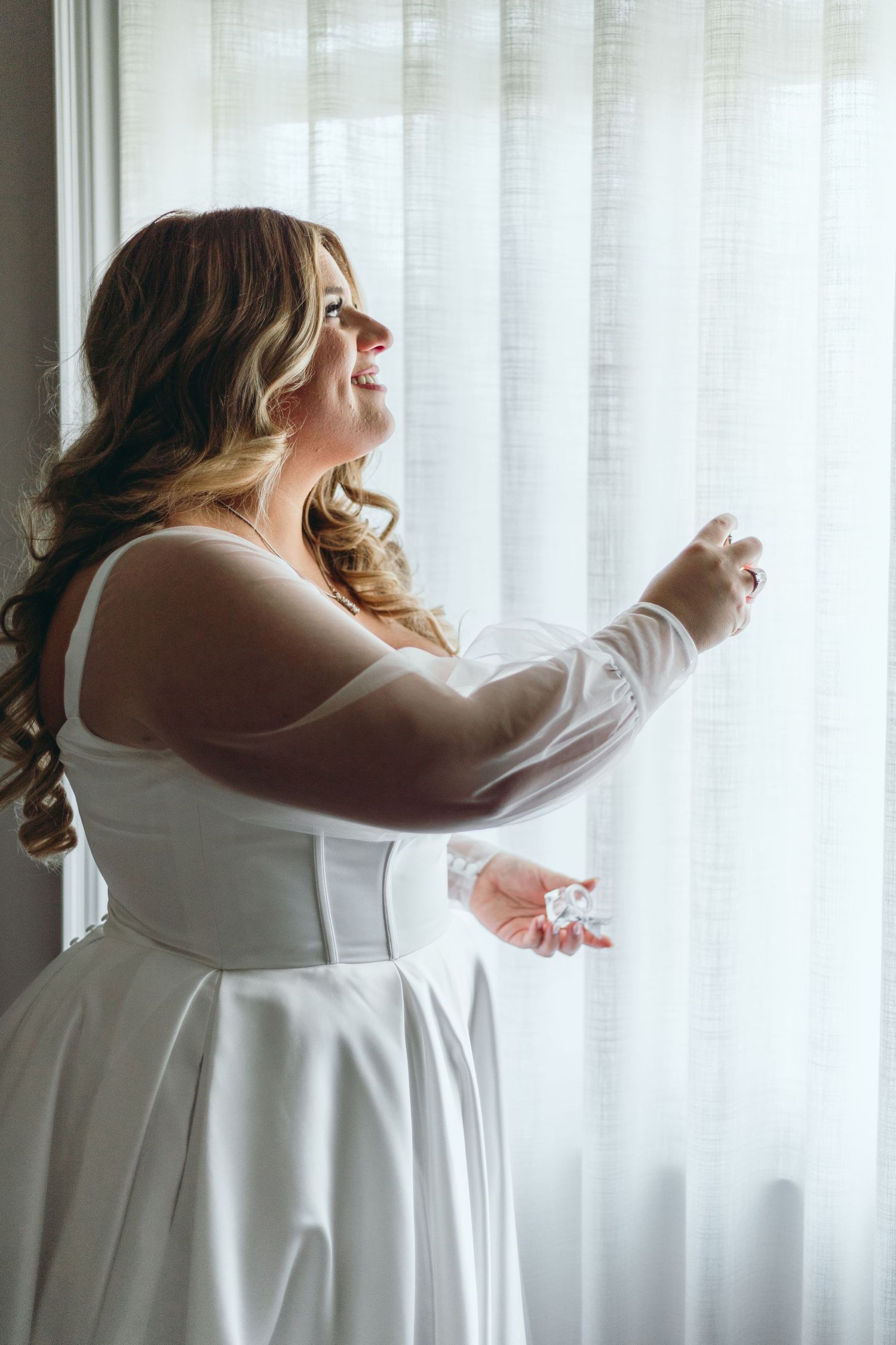 Bride in white dress twirls against sheer curtained window.