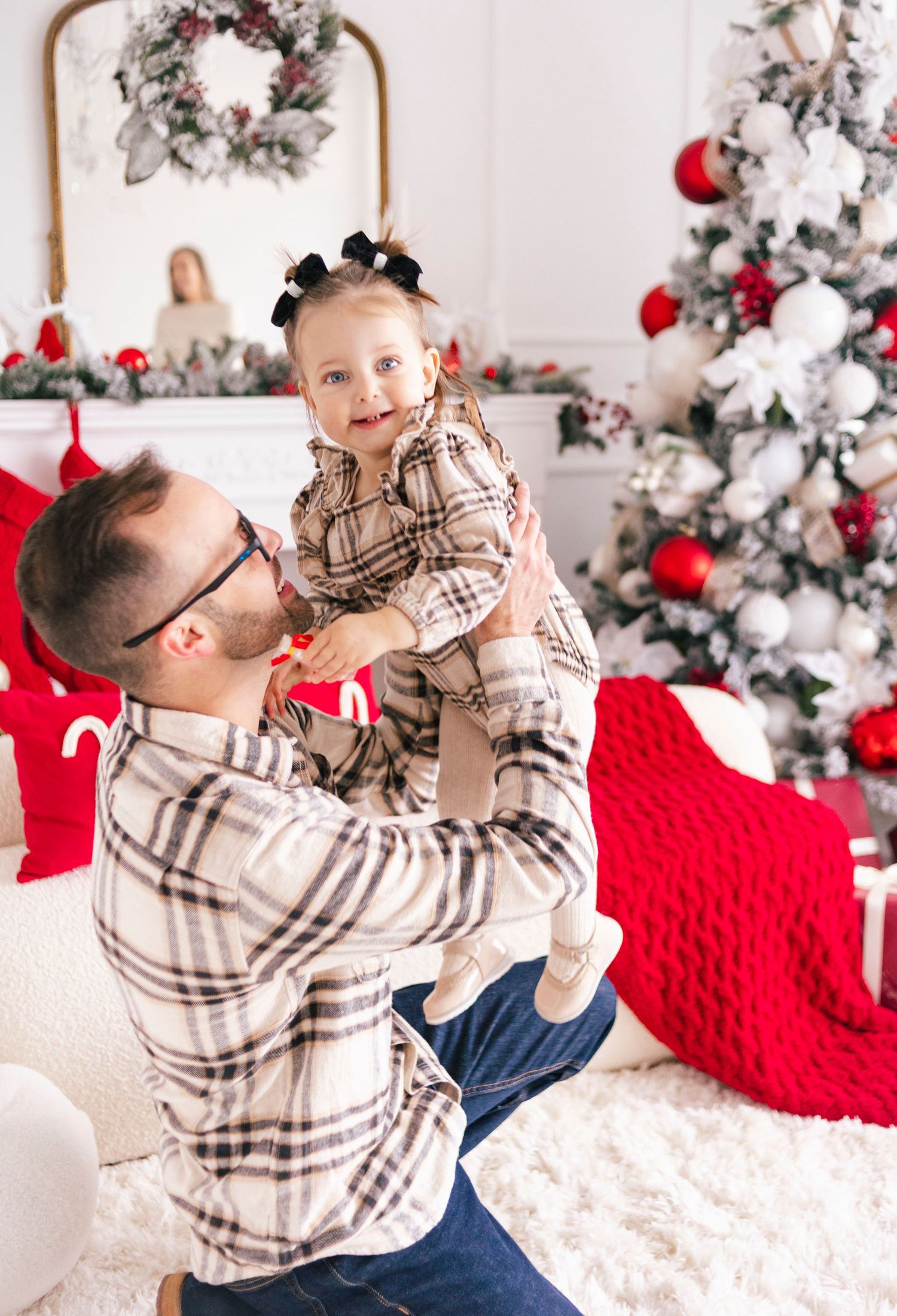 Father lifting smiling baby girl in matching plaid outfits near decorated Christmas tree with red and white ornaments.