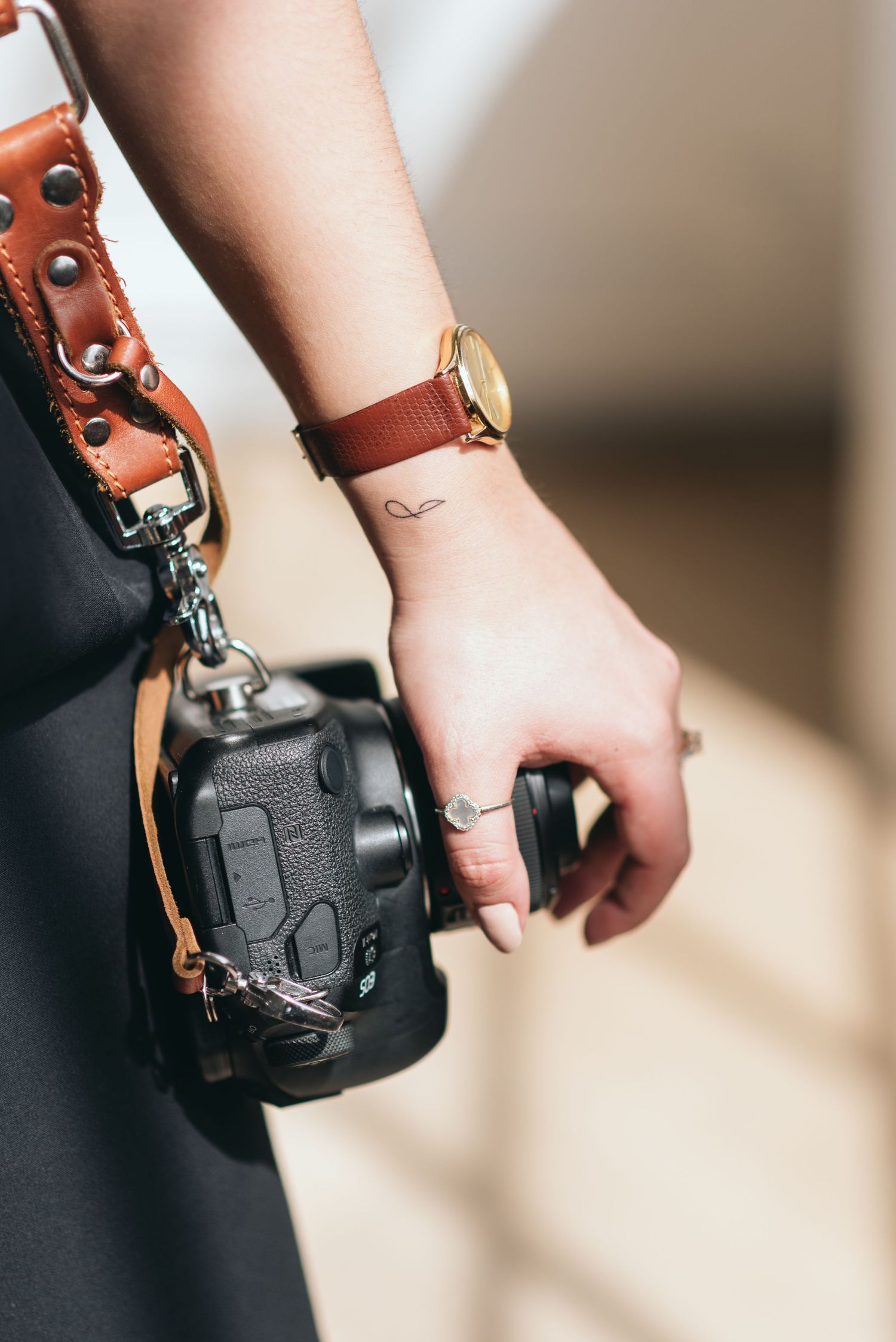 A hand wearing a brown leather camera strap holds a black DSLR camera against a blurred background.
