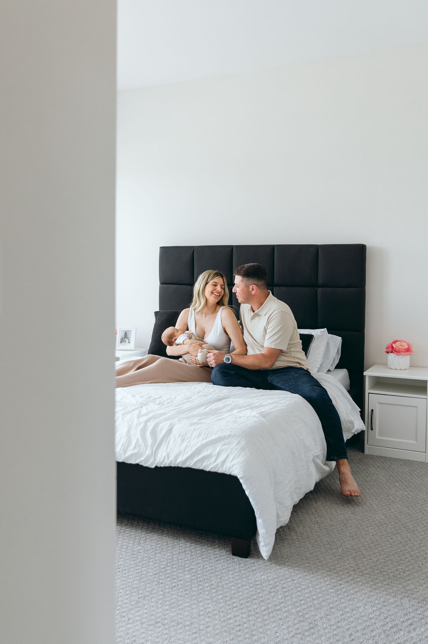 Family portrait on a modern bed with black headboard and white bedding in a bright room.