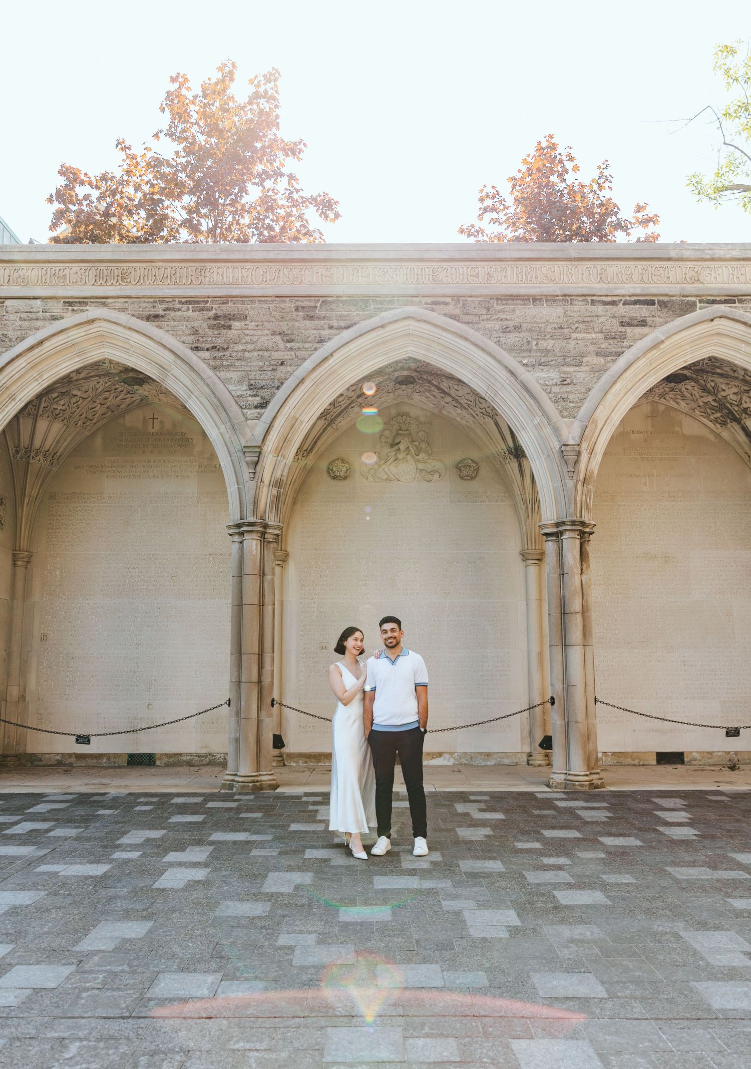 Couple in white attire poses against a stone archway with elegant architectural details.