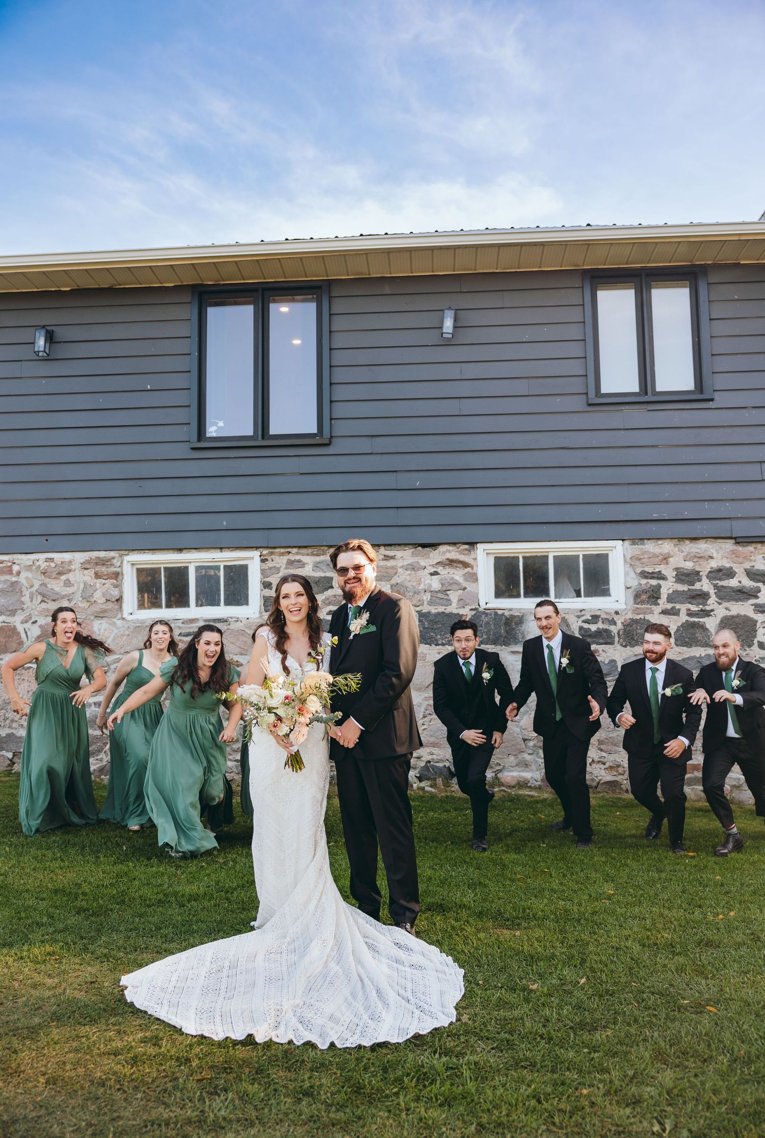 Wedding party poses playfully in a line on grass with stone wall and grey building backdrop.
