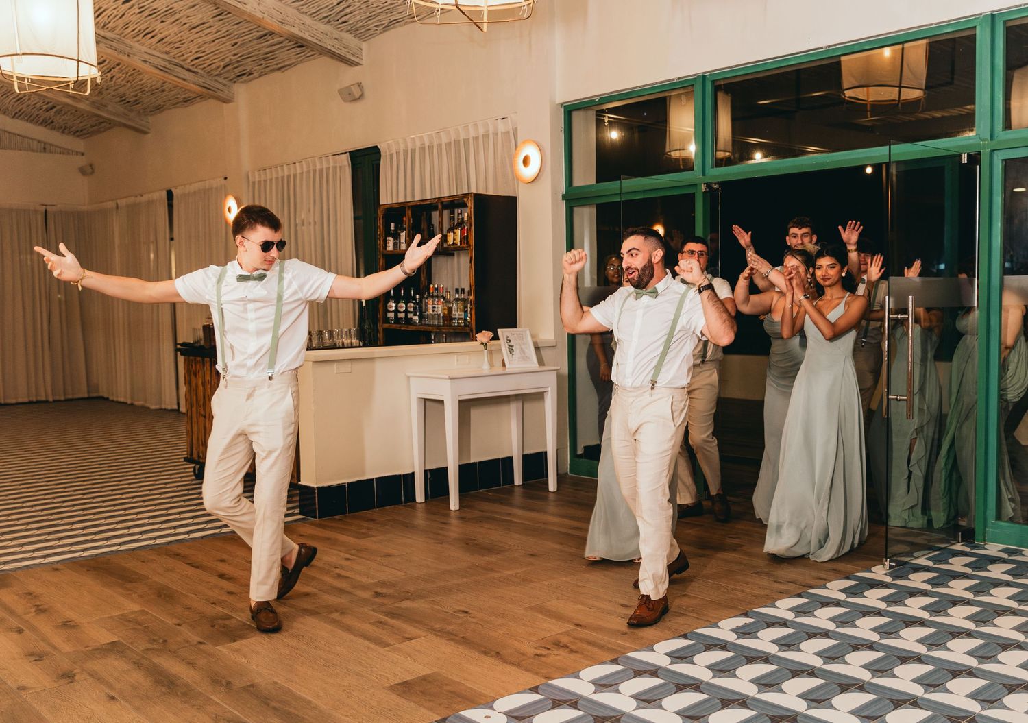 Wedding party performs synchronized dance moves in a reception venue with wooden floors.