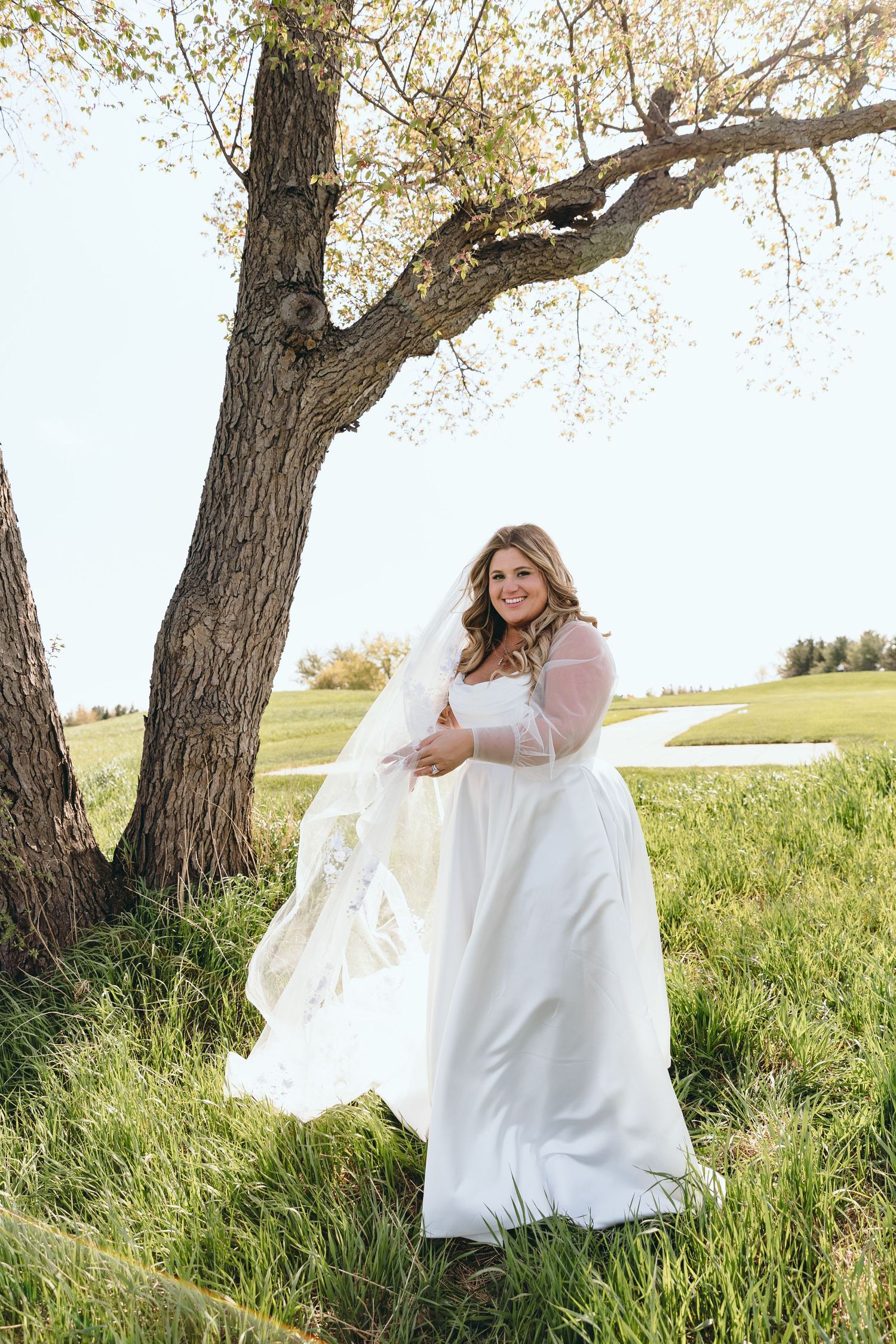 A bride in a flowing white dress poses under a tree in a grassy field during a sunny outdoor photoshoot.