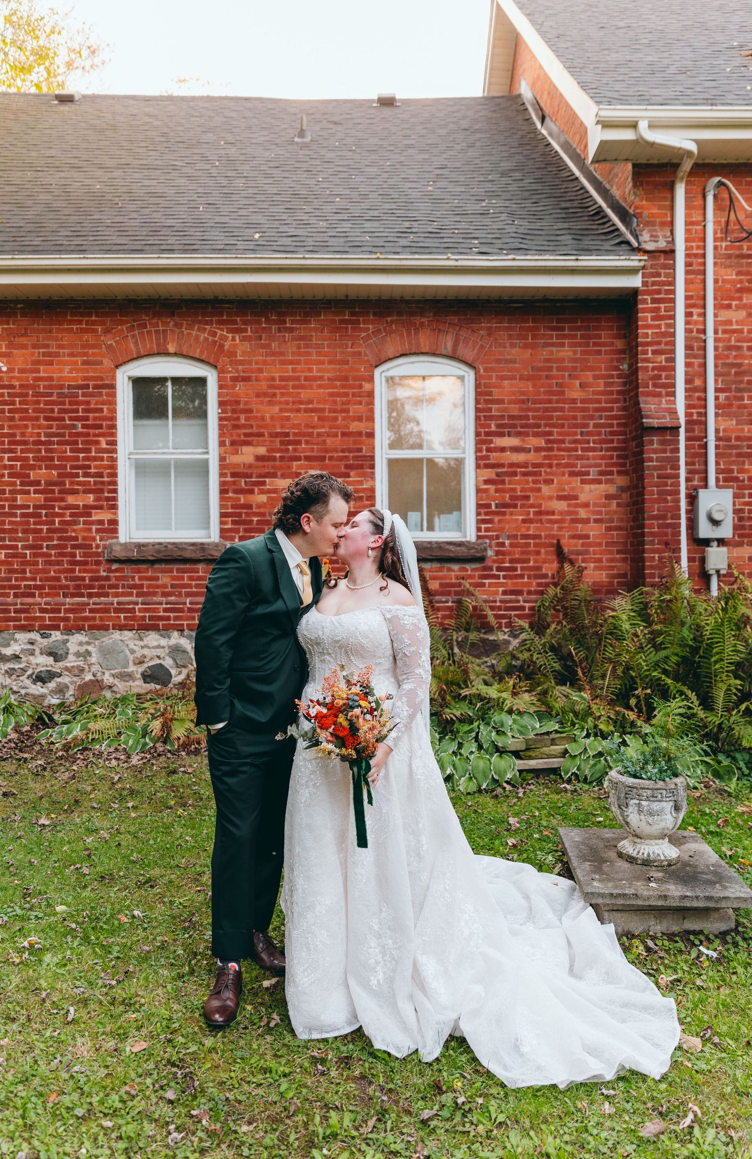 A couple in wedding attire poses romantically against a brick building backdrop.