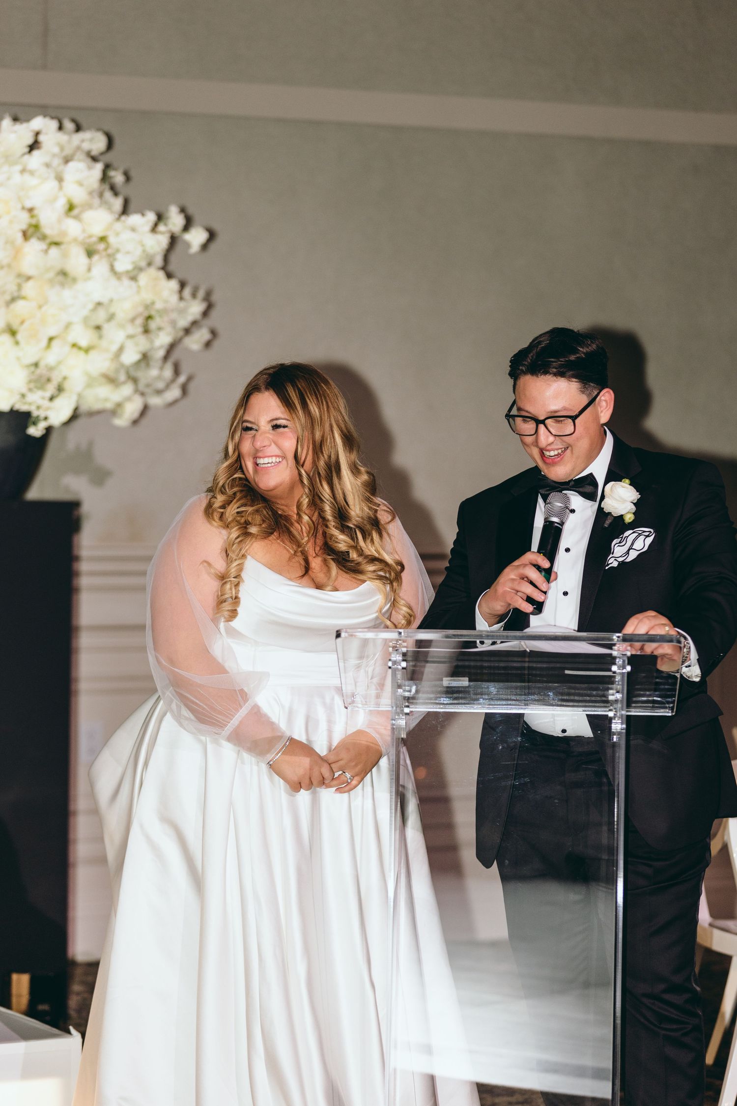 Couple shares moment at clear podium during wedding reception speech.