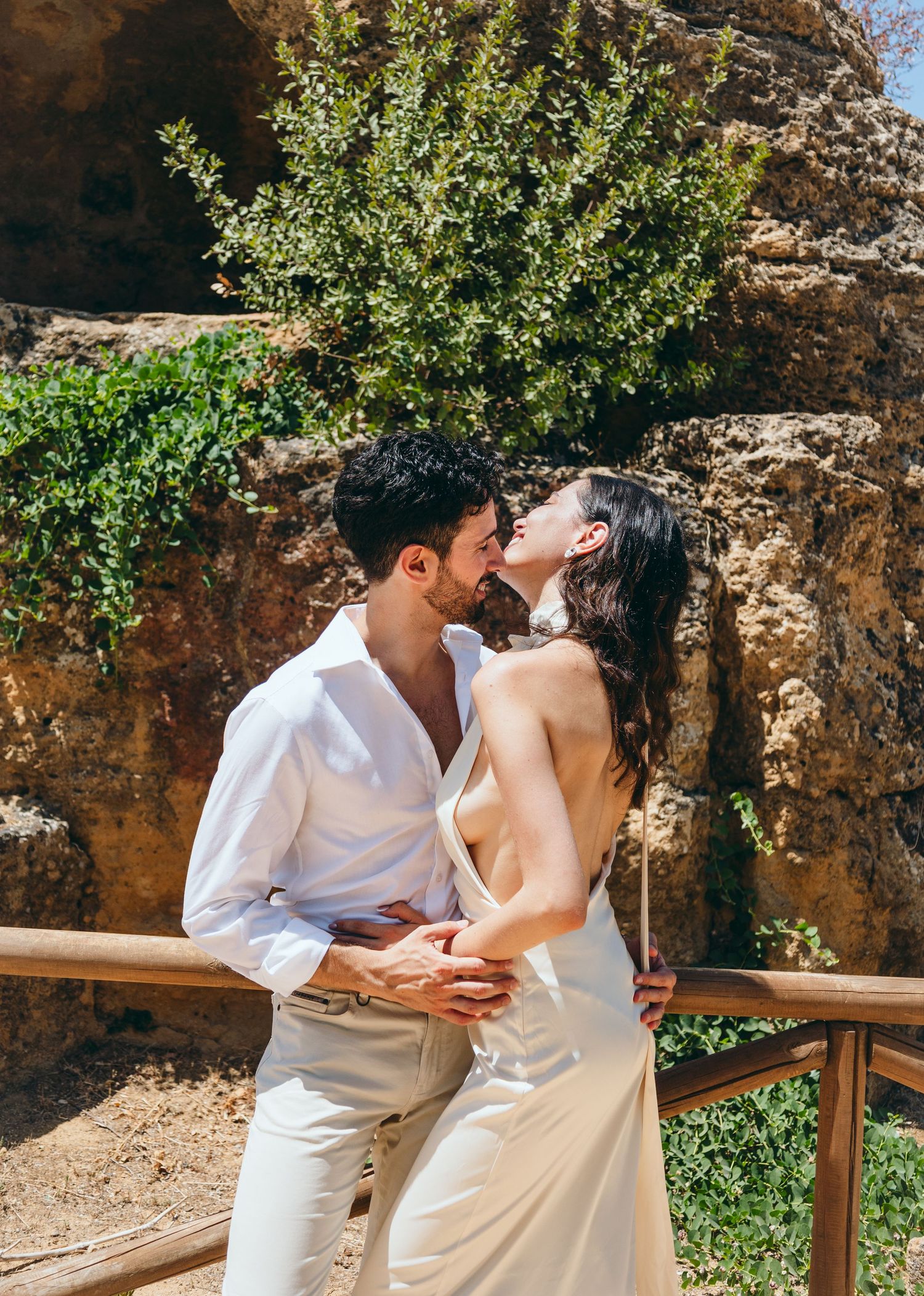 A couple embraces on a rustic pathway with green foliage and stone walls in the background.