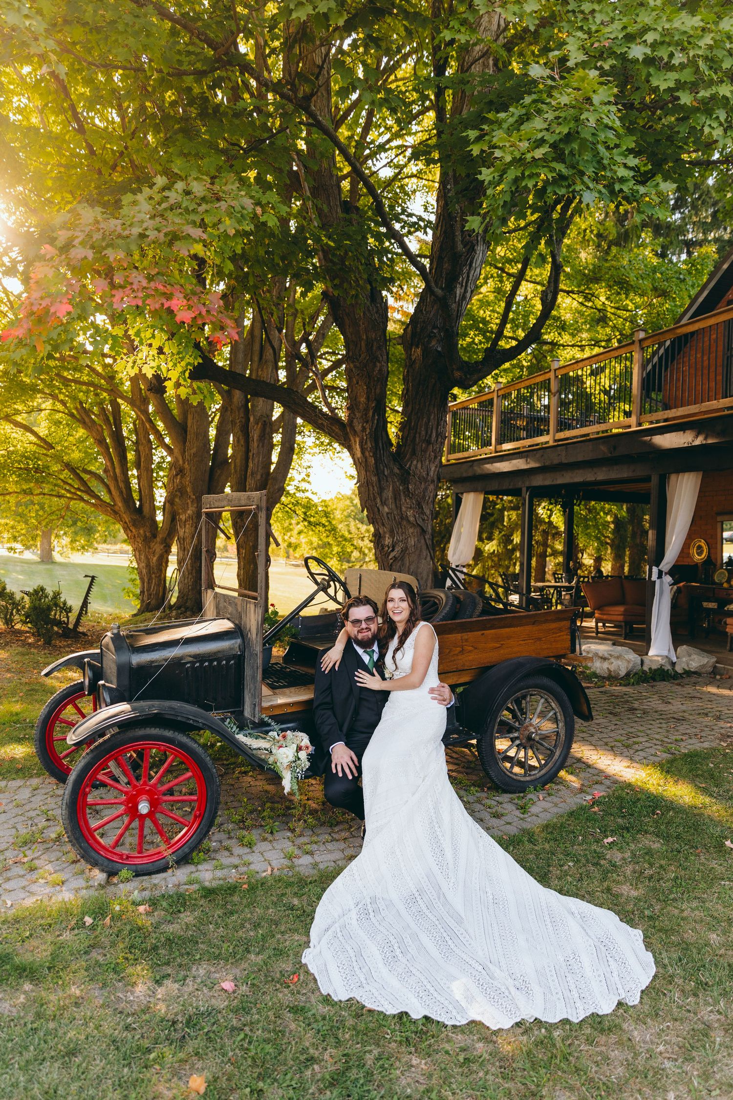 A vintage car wedding portrait with golden sunlight filtering through trees near a rustic building.