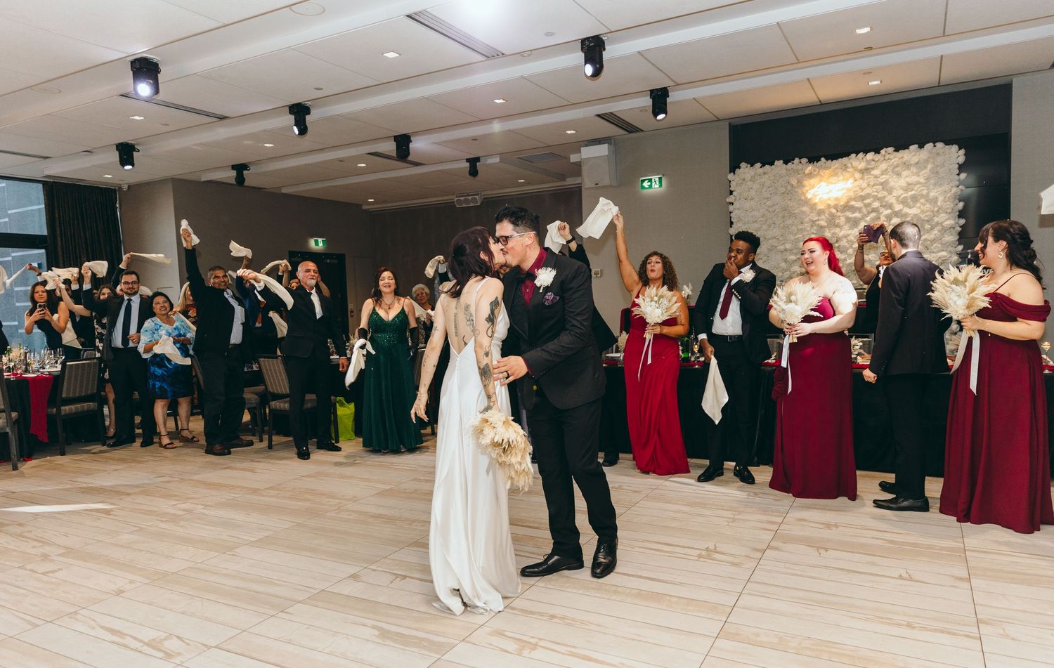 Wedding guests dancing together on a light wooden dance floor at an indoor reception venue.
