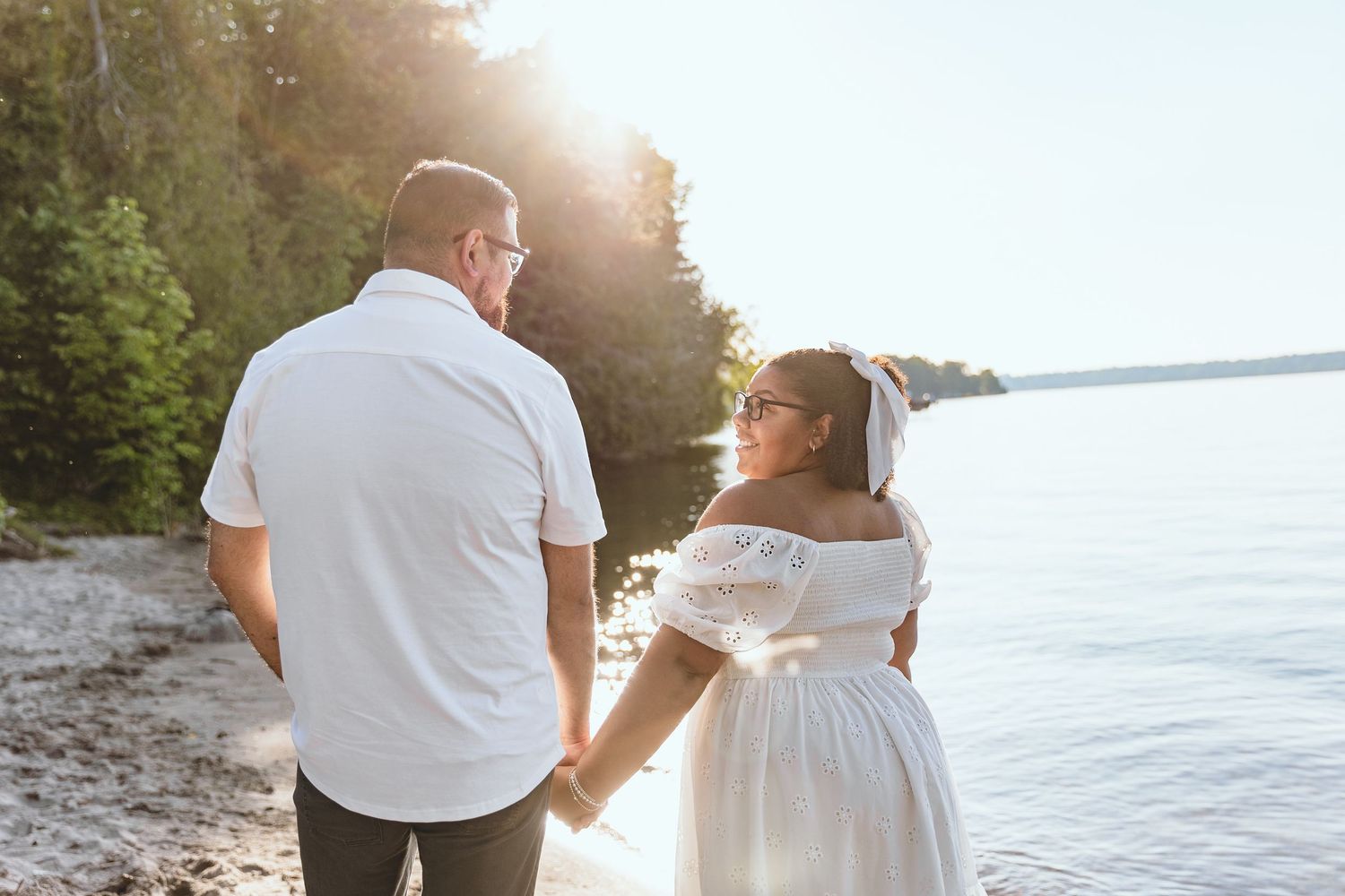Couple walking along shoreline at golden hour holding hands in white outfits.