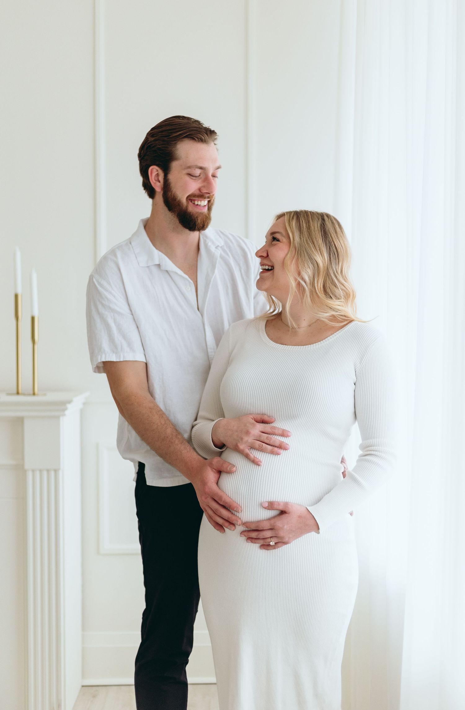 Joyful maternity session with couple in white sharing laughs against bright background.