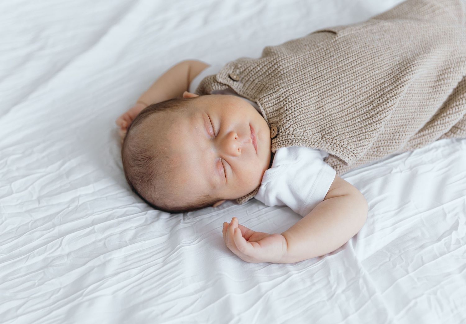 Infant sleeping soundly on white bedding while holding knitted teddy bear toy.