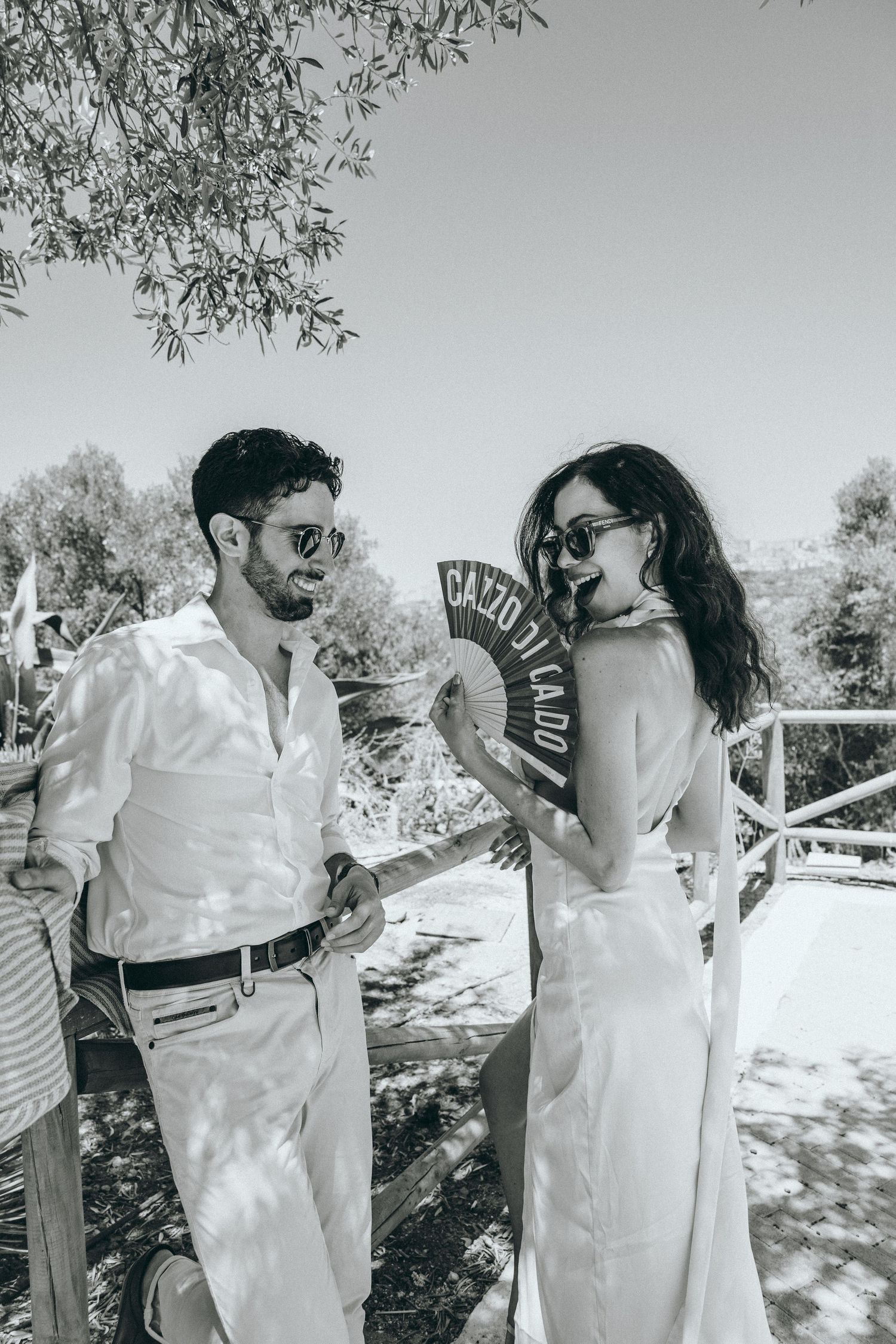Playful couple in white outfits share drinks during sunny outdoor photoshoot.