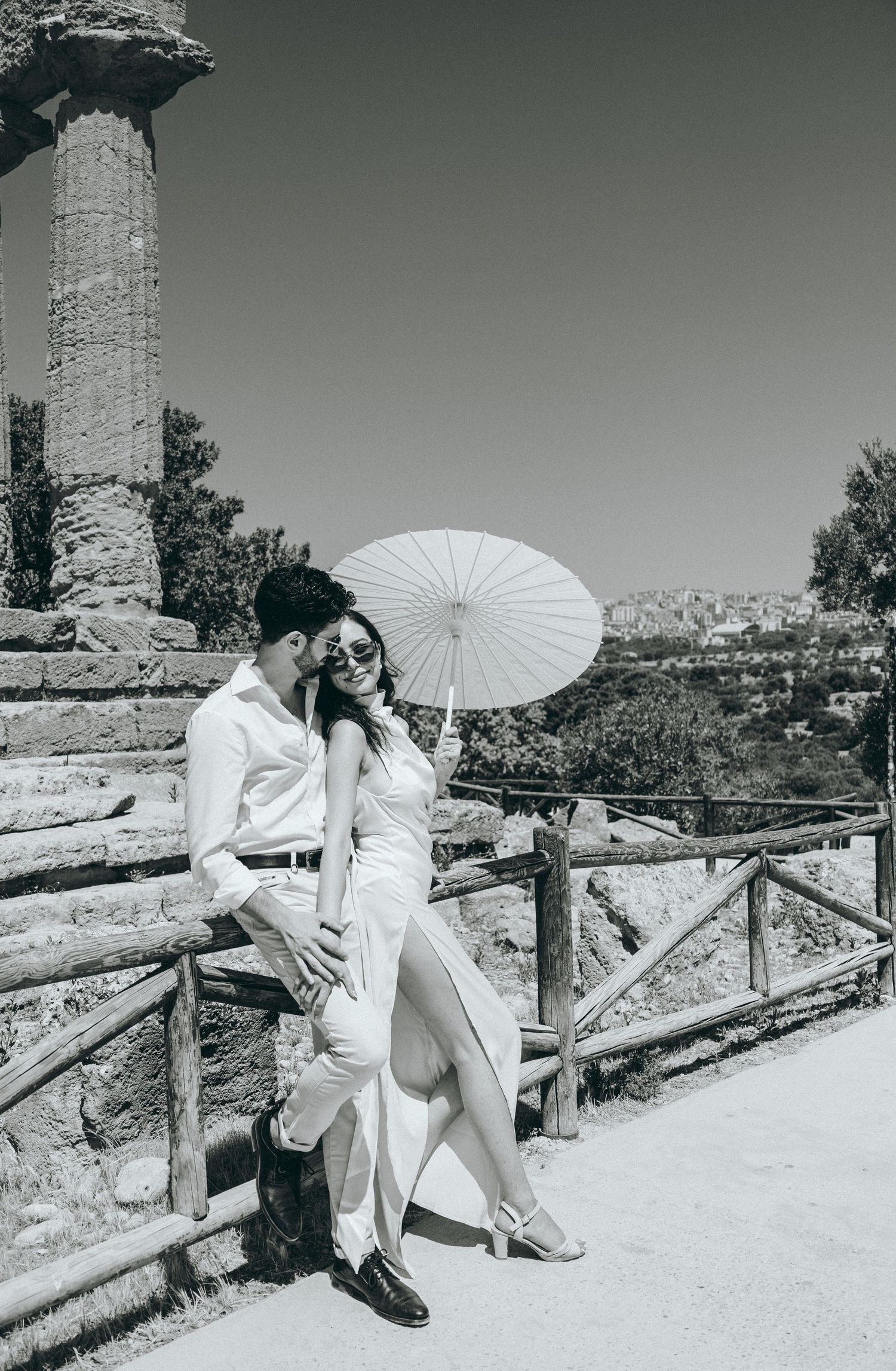 Stylish couple with white parasol pose along wooden fence at historic site.