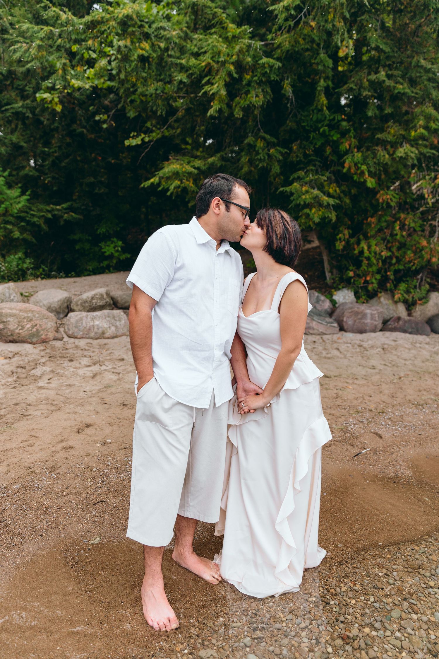 Couple in white clothing sharing an intimate moment on a beach with trees in background.