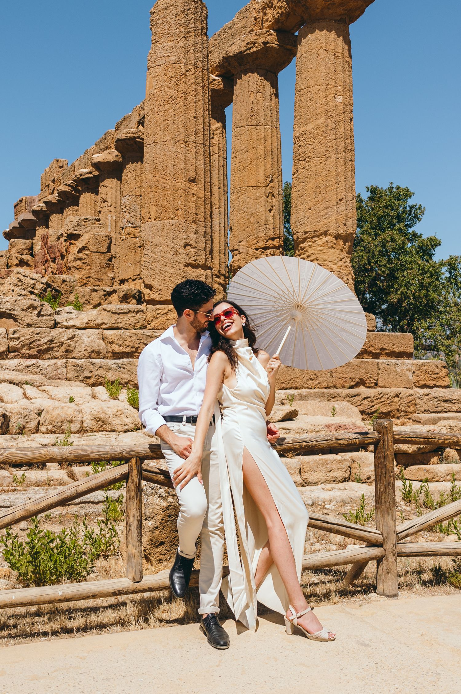 Elegant couple with parasol pose near ancient Greek temple ruins on sunny day in Sicily.
