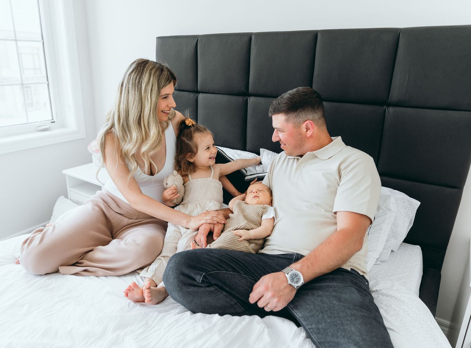 Family relaxing together on black upholstered bed in bright white room.