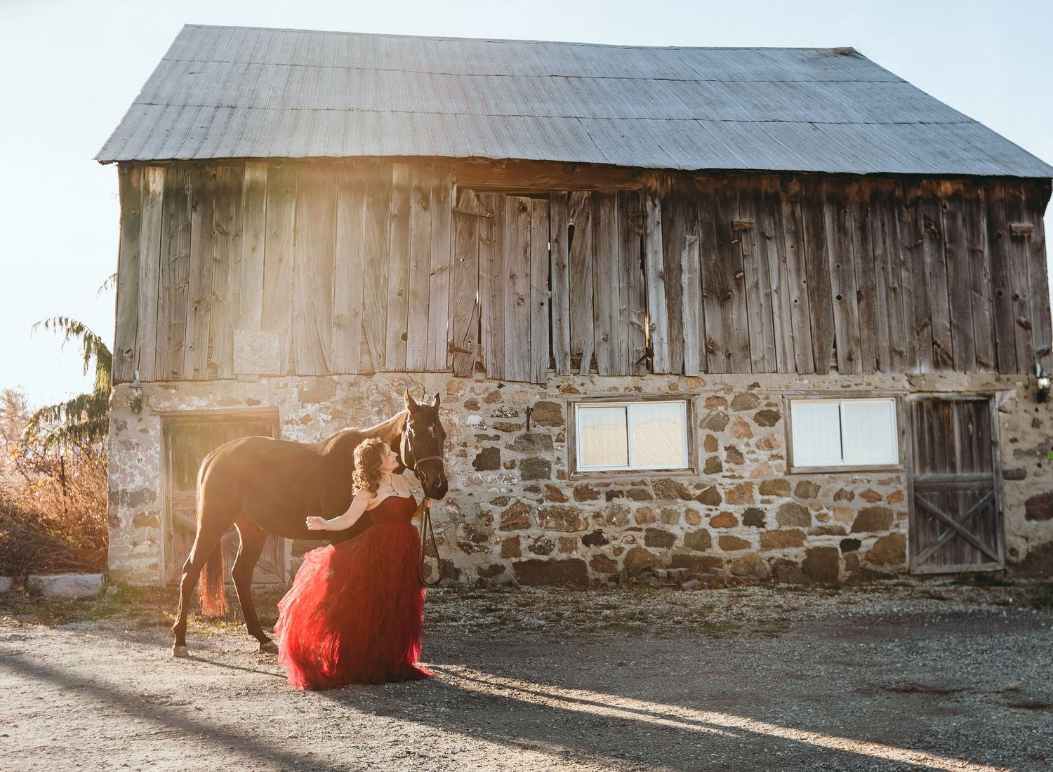 A person in a flowing red dress stands beside a horse against the backdrop of a stone and wood barn.