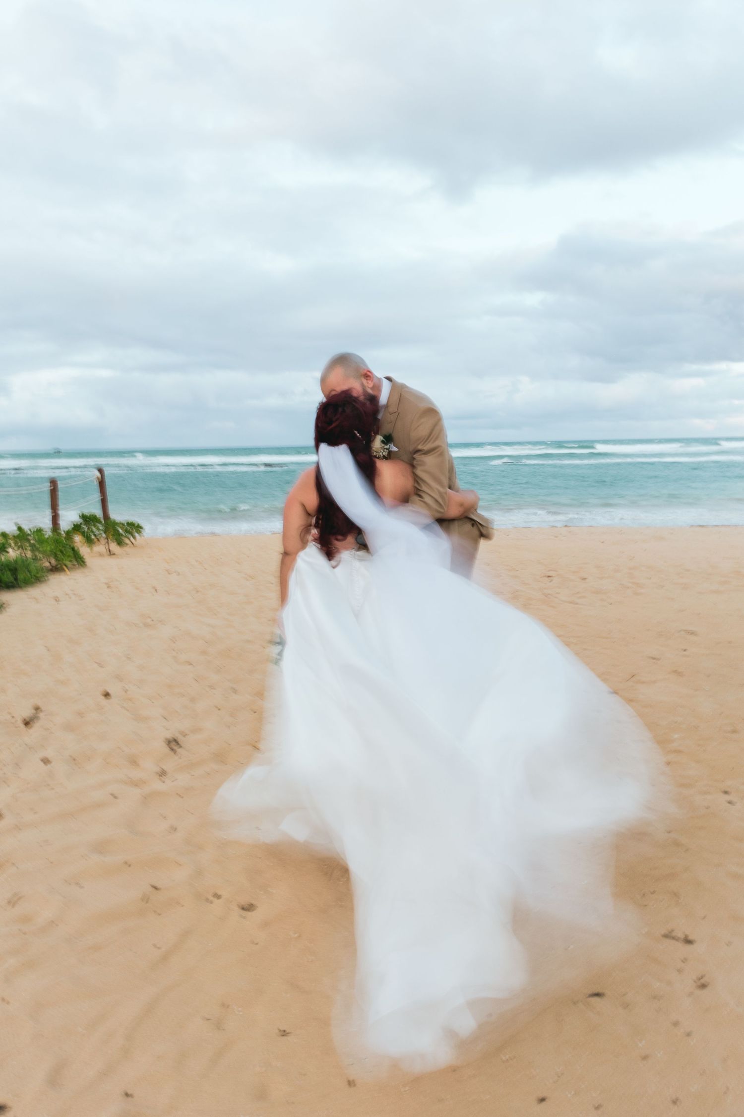 Newlyweds share an intimate moment on a tropical beach with crashing waves in the background.