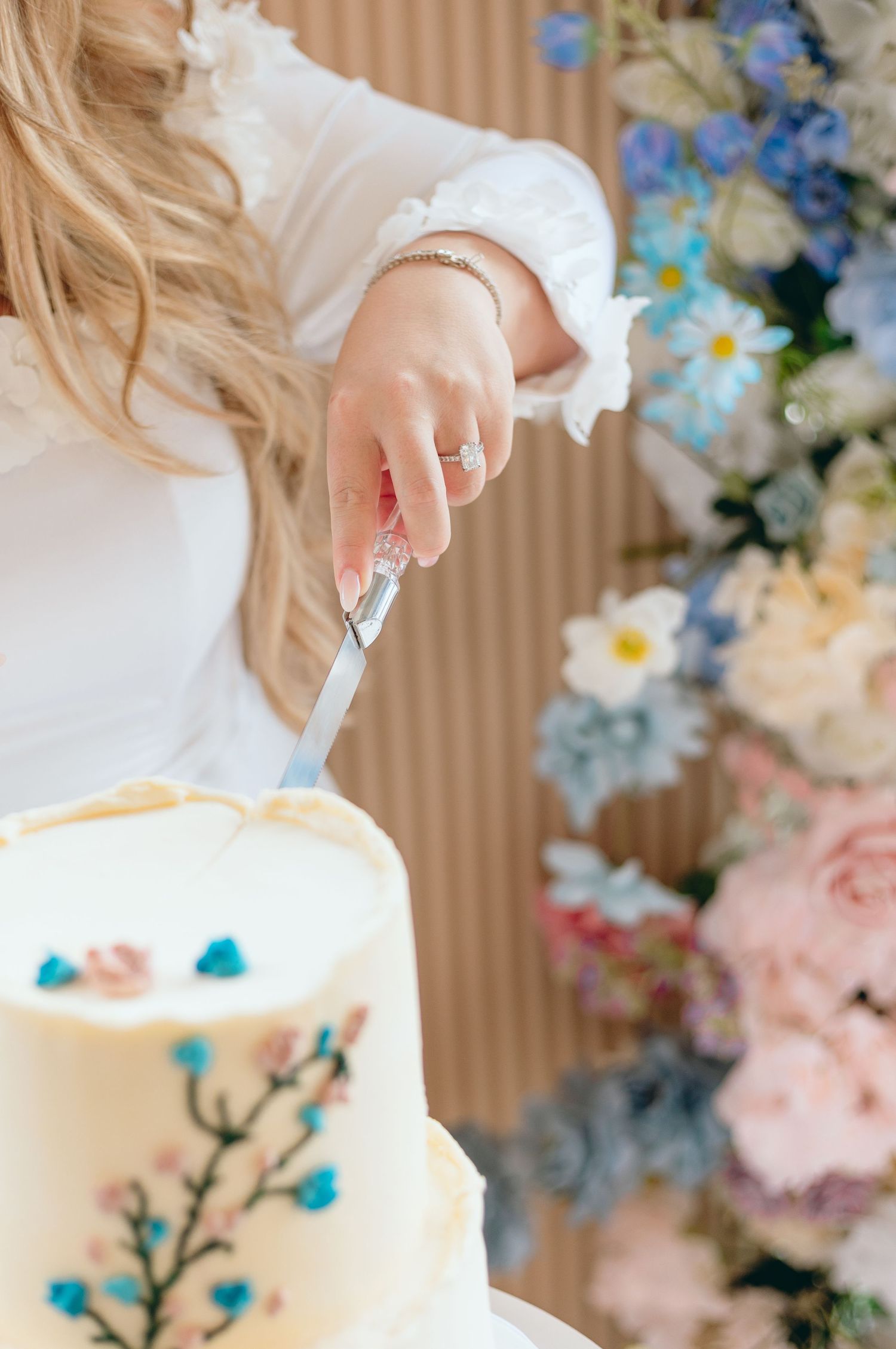A hand delicately pipes blue floral decorations onto a white wedding cake with multiple tiers.
