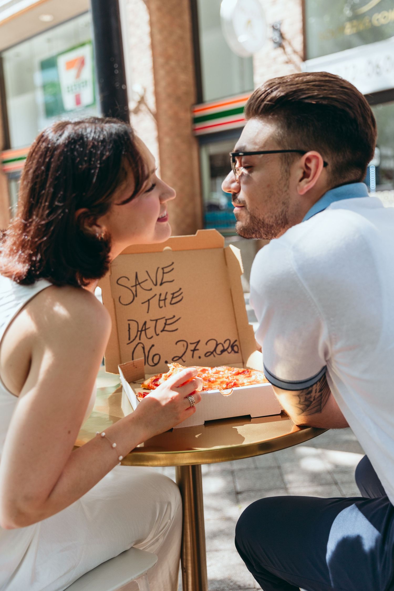 A couple shares pizza while holding save the date signs.