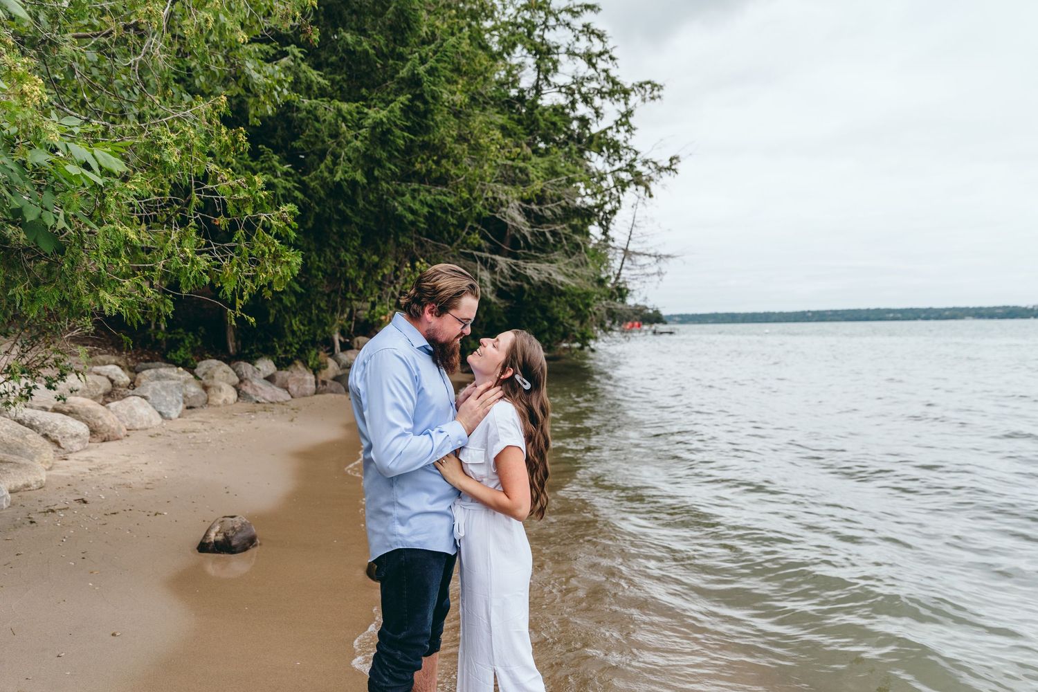 Couple embraces on a scenic beach shoreline with trees and water in the background.