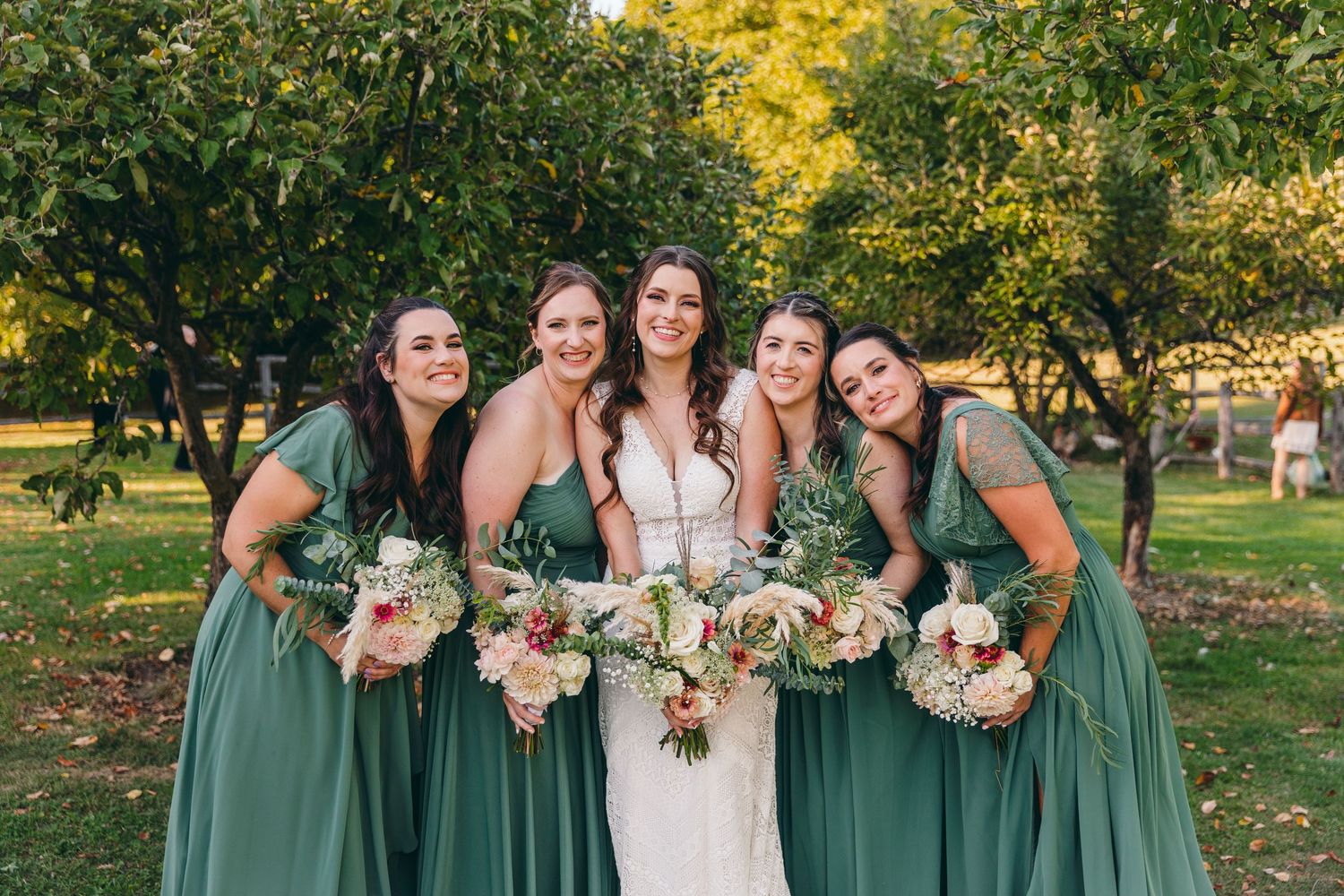 Bridesmaids in sage green dresses pose together outdoors in autumn setting.