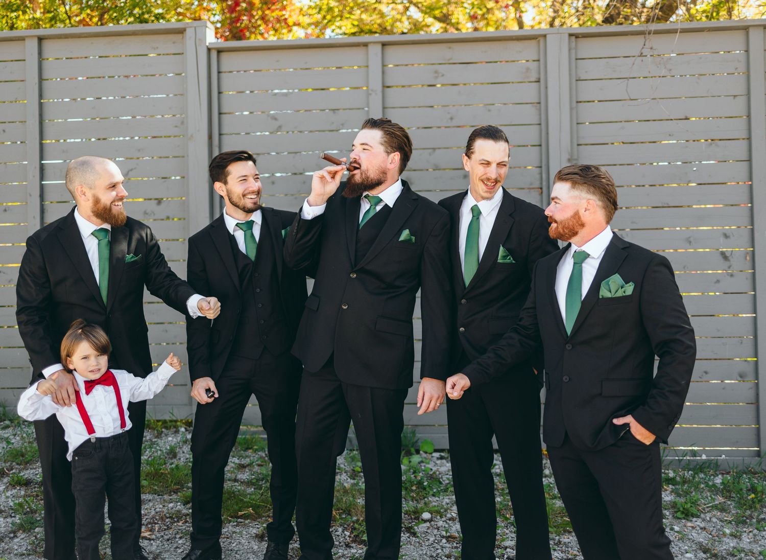Groomsmen in black suits and green ties lined up against a modern grey fence.