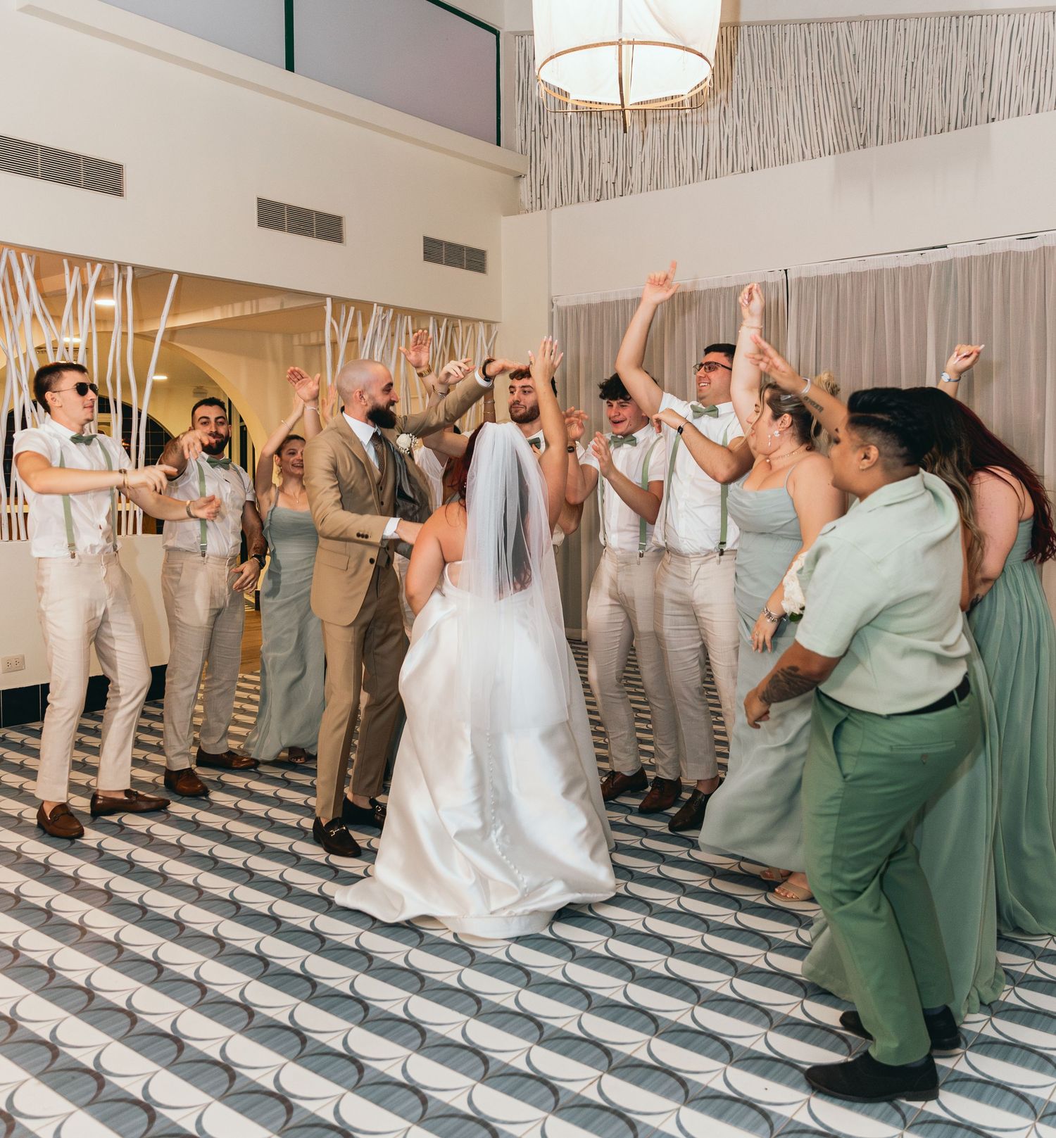Wedding party in sage green dresses and white attire dances together on a patterned floor.