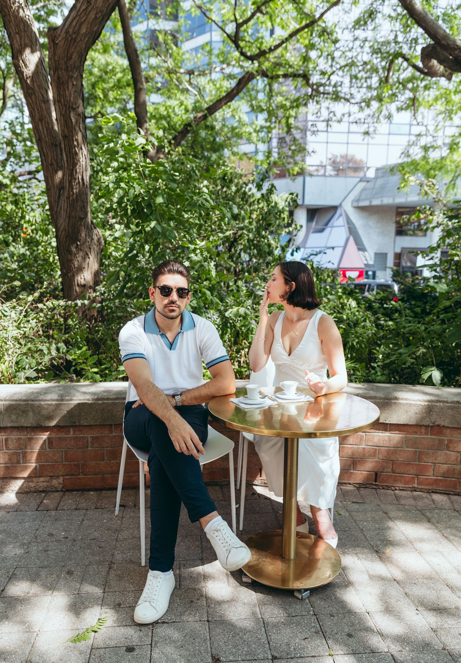 A casual outdoor dining scene with people seated at a metal table under leafy trees.