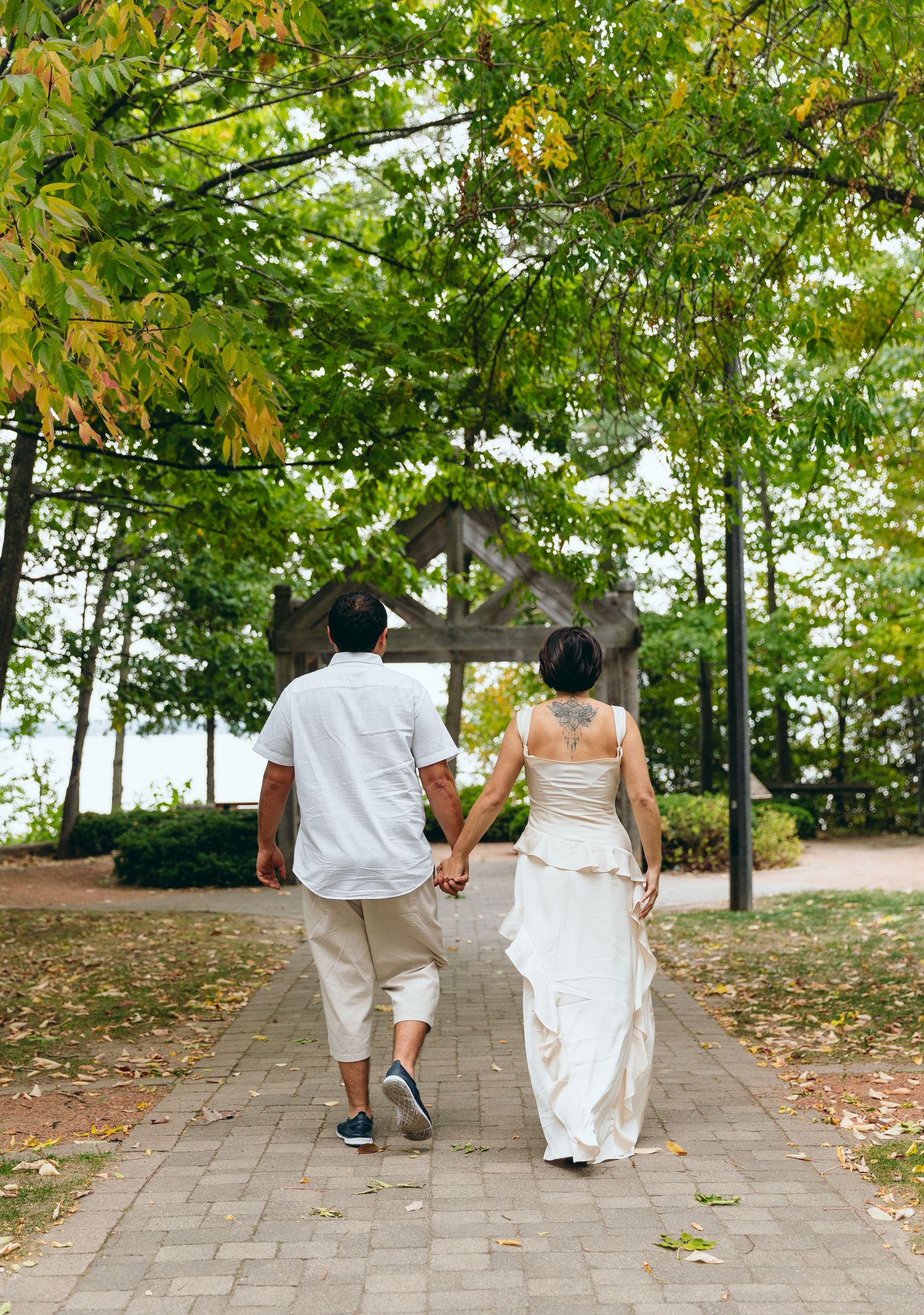 A couple in white attire walking hand in hand down a tree-lined path towards a gazebo.