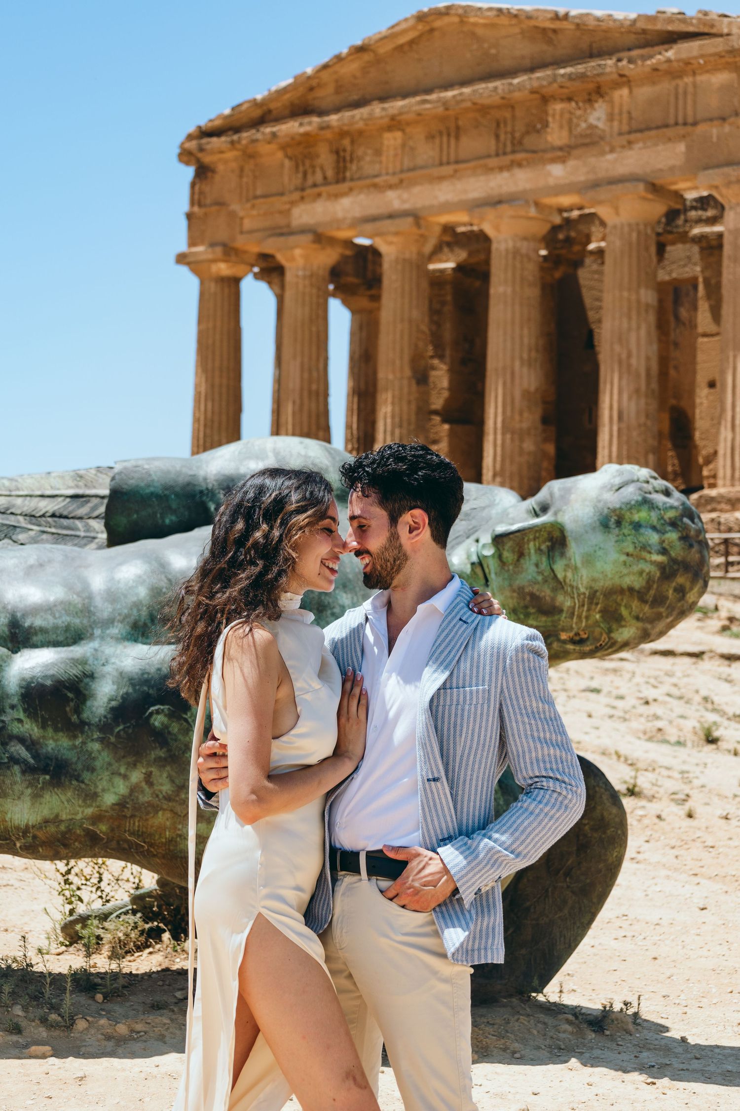 Couple embracing in front of historic Greek temple columns under bright blue sky.