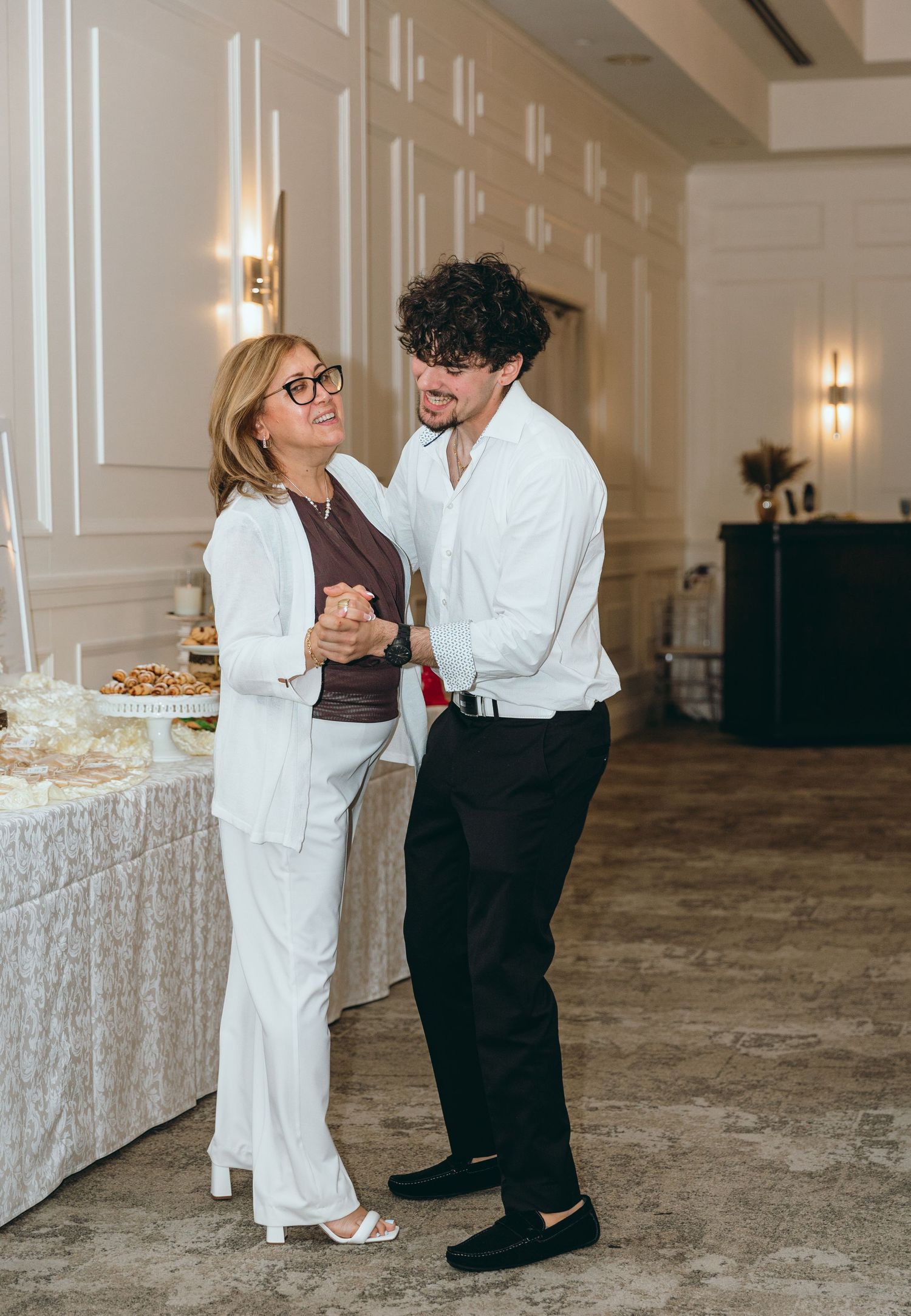 A couple shares an intimate dance moment in an elegant reception hall with white paneled walls.